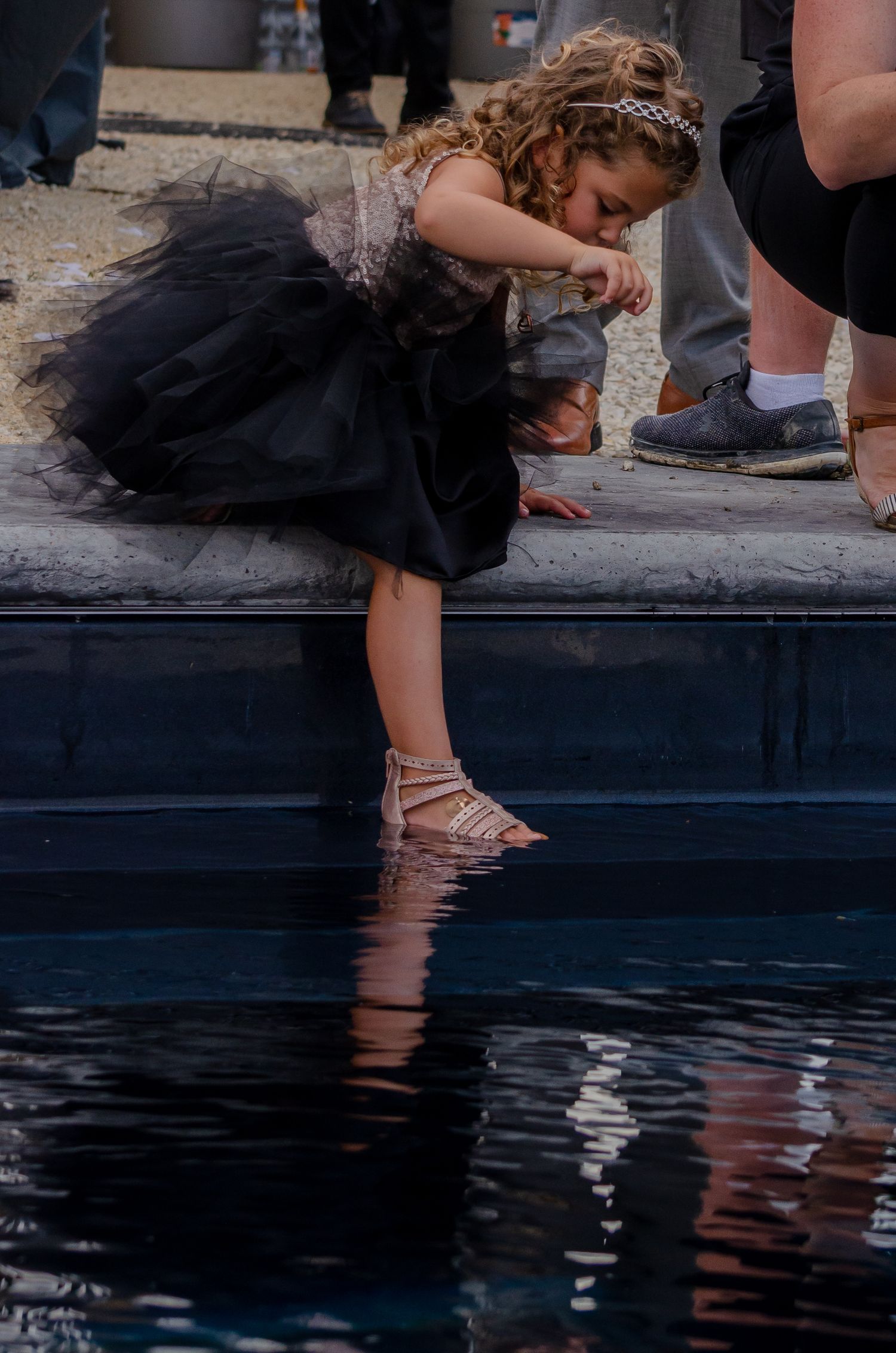 Young girl in black tulle dress dips her feet into a reflective pool, bending forward curiously.