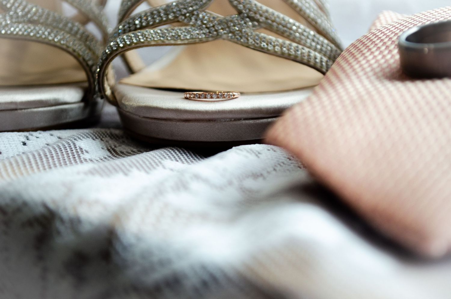 Elegant silver strappy heels with rhinestones beside a pink tie and wedding rings on a textured cloth.