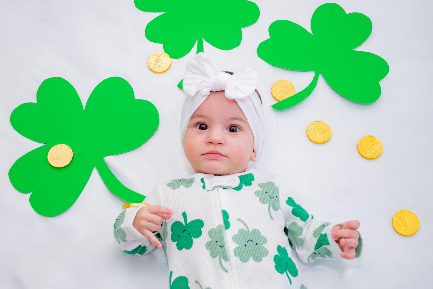 Baby in shamrock outfit lying with green clover decorations and gold coins for St. Patrick's Day.