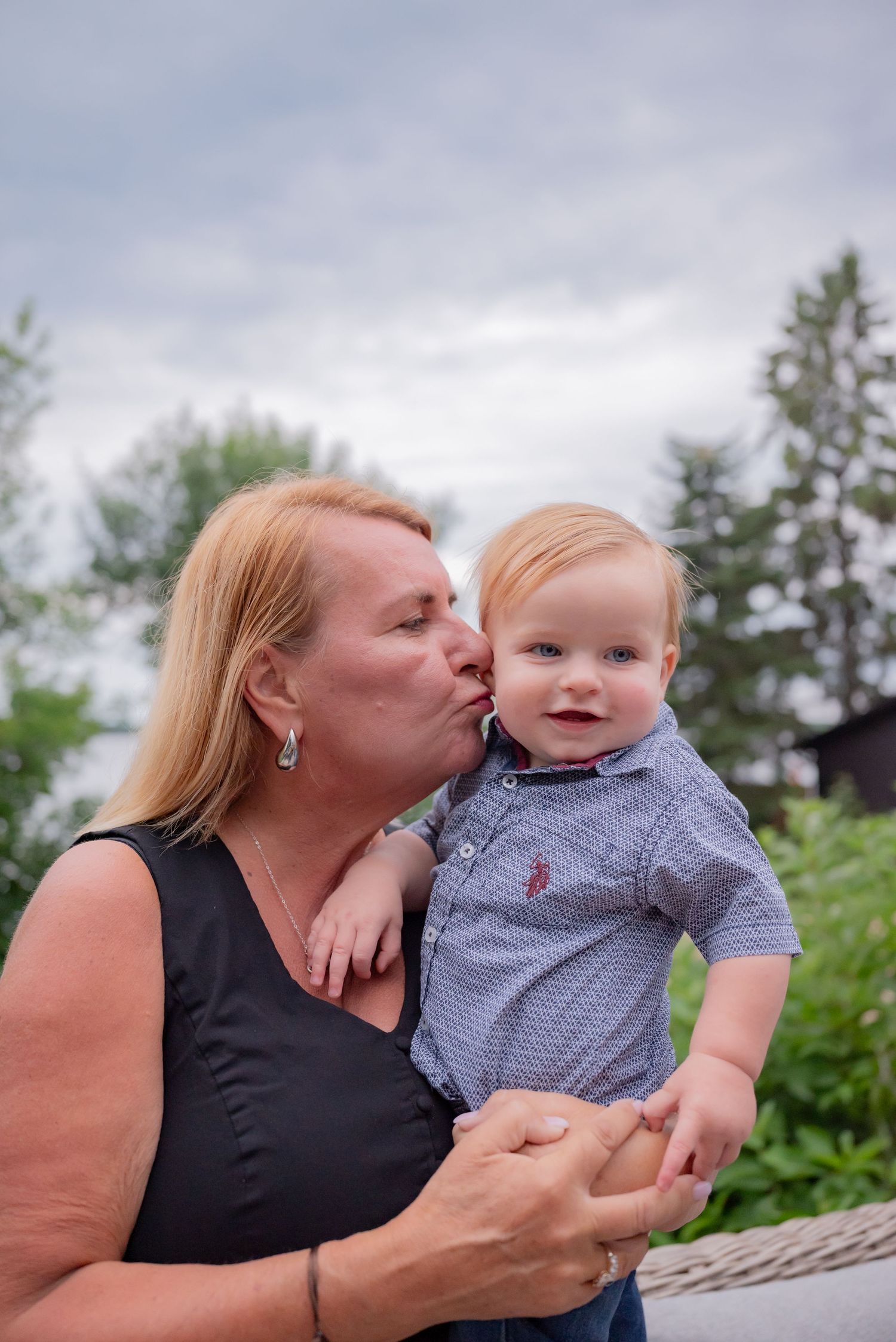 A grandmother with blonde hair kisses a happy blue-eyed baby boy on the cheek outdoors.