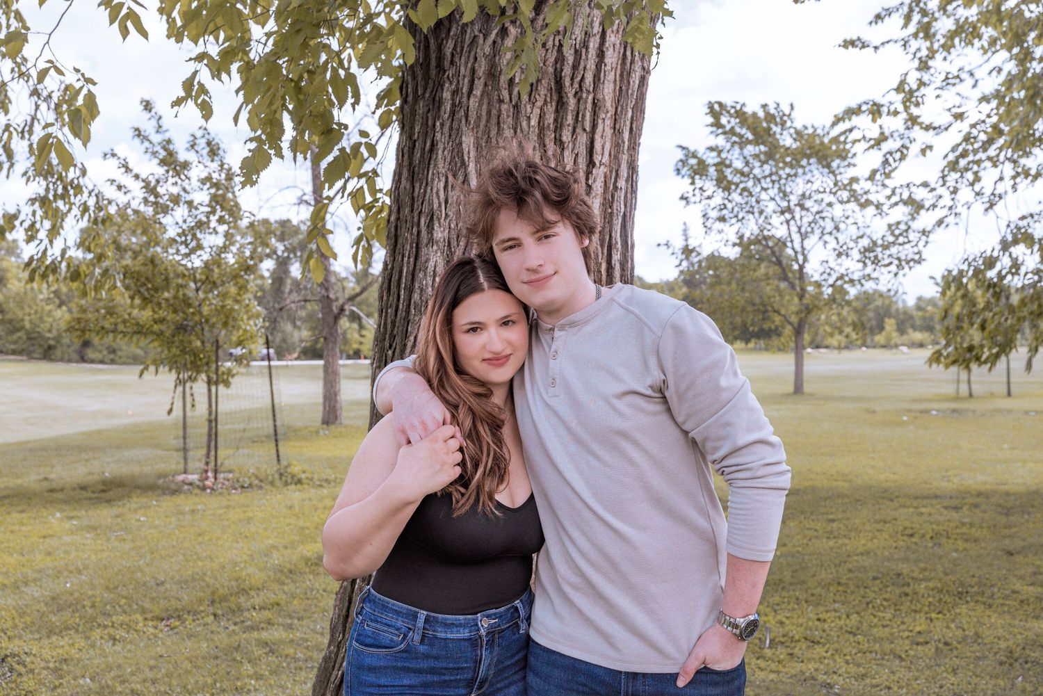Two teenagers posing together in front of a large tree in a green park on a cloudy day.