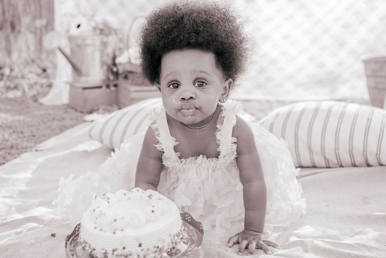 Black and white photo of a baby girl in a white dress sitting near a birthday cake during a cake smash photoshoot.