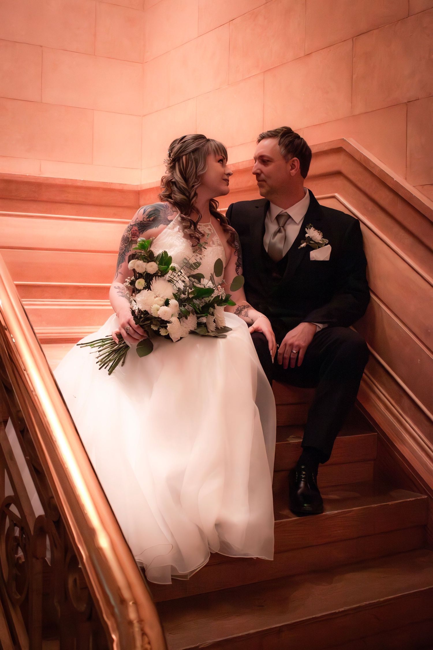 Bride and groom sit together on elegant staircase, sharing a romantic gaze, bride holding white floral bouquet.