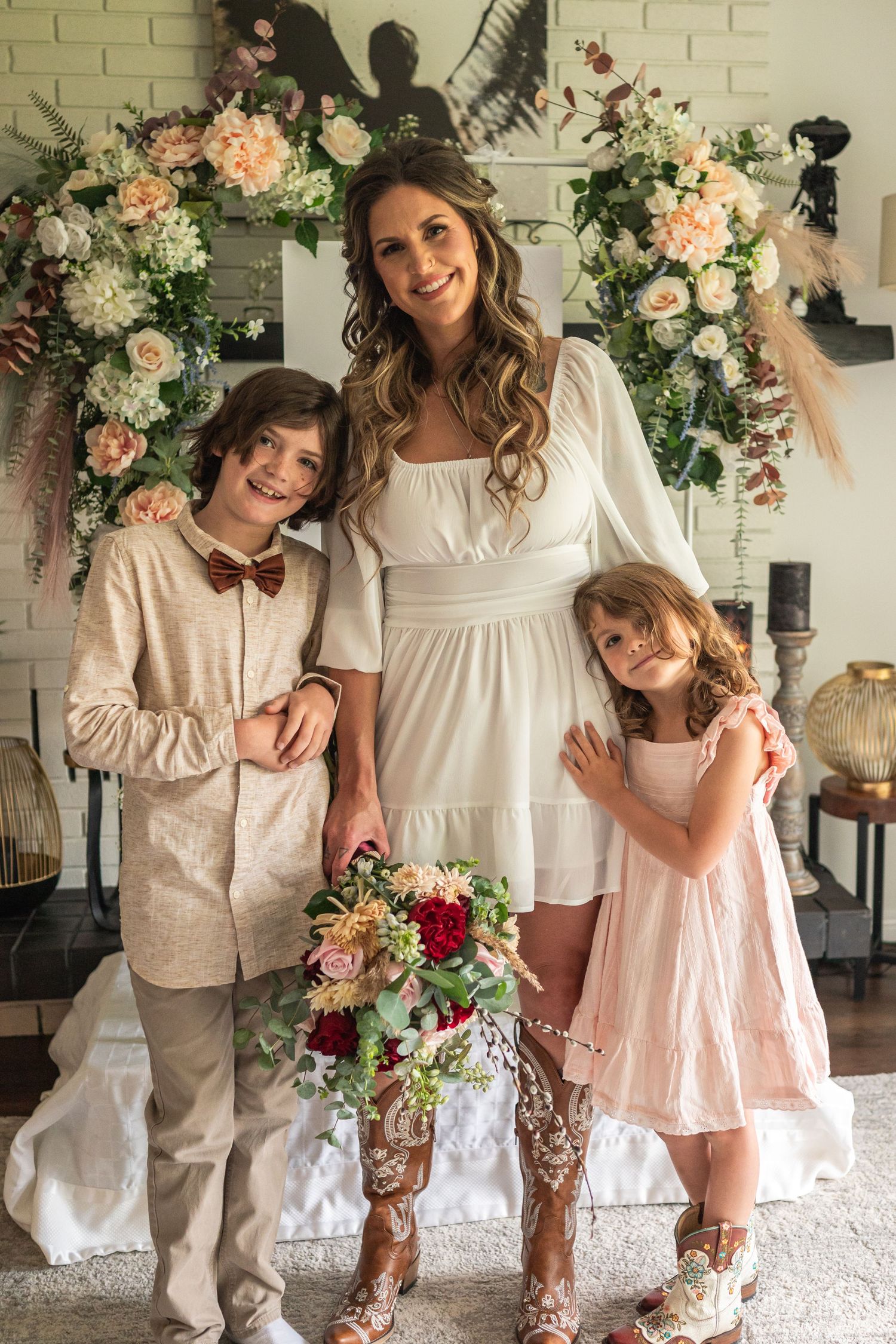 Bride in white dress poses with two children before floral arch backdrop at wedding ceremony.