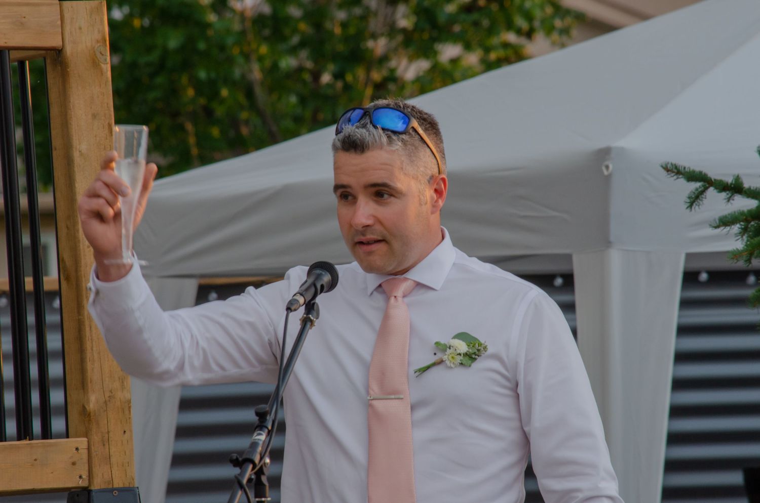 Man in white shirt and pink tie raises champagne glass while giving a toast at an outdoor wedding celebration.