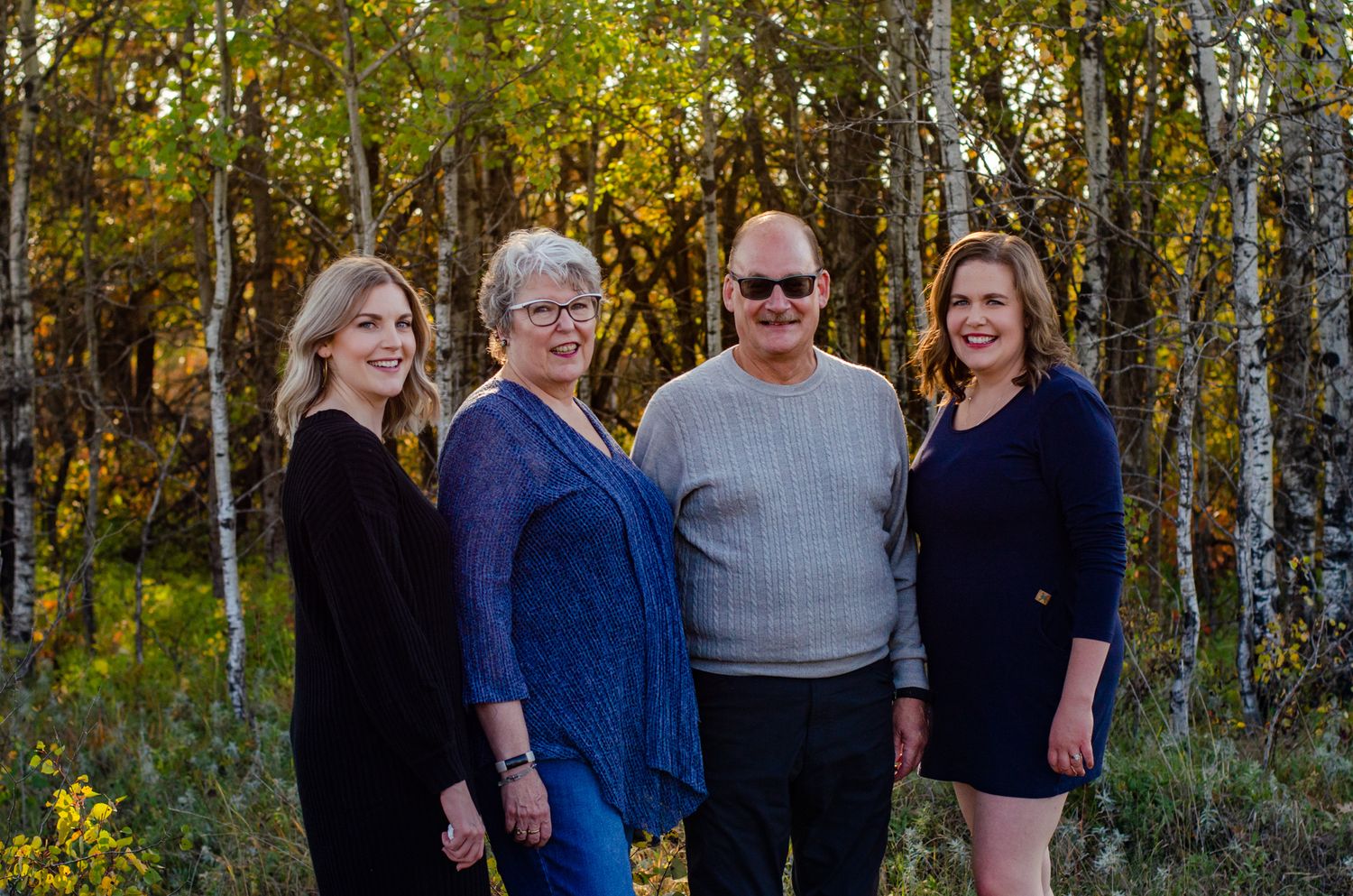 Family of four smiling outdoors among autumn birch trees in a warm fall nature portrait.