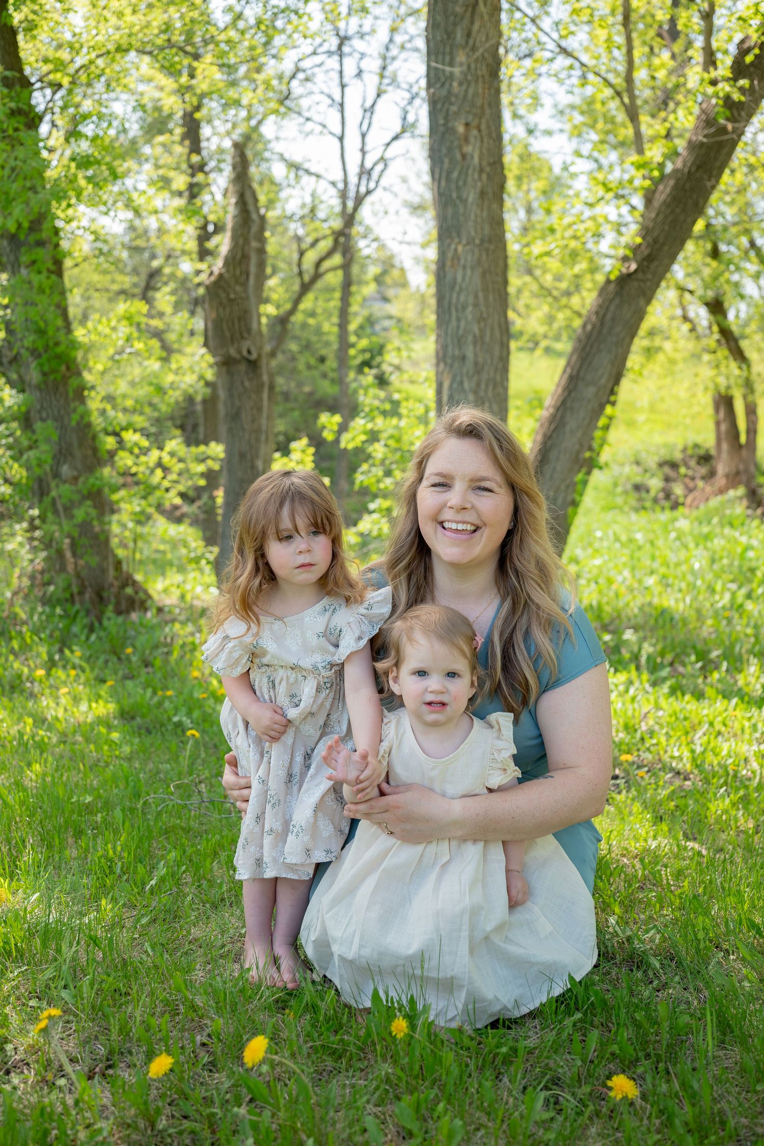 A smiling woman kneels outdoors with two young girls in white dresses, surrounded by lush green trees and dandelions.