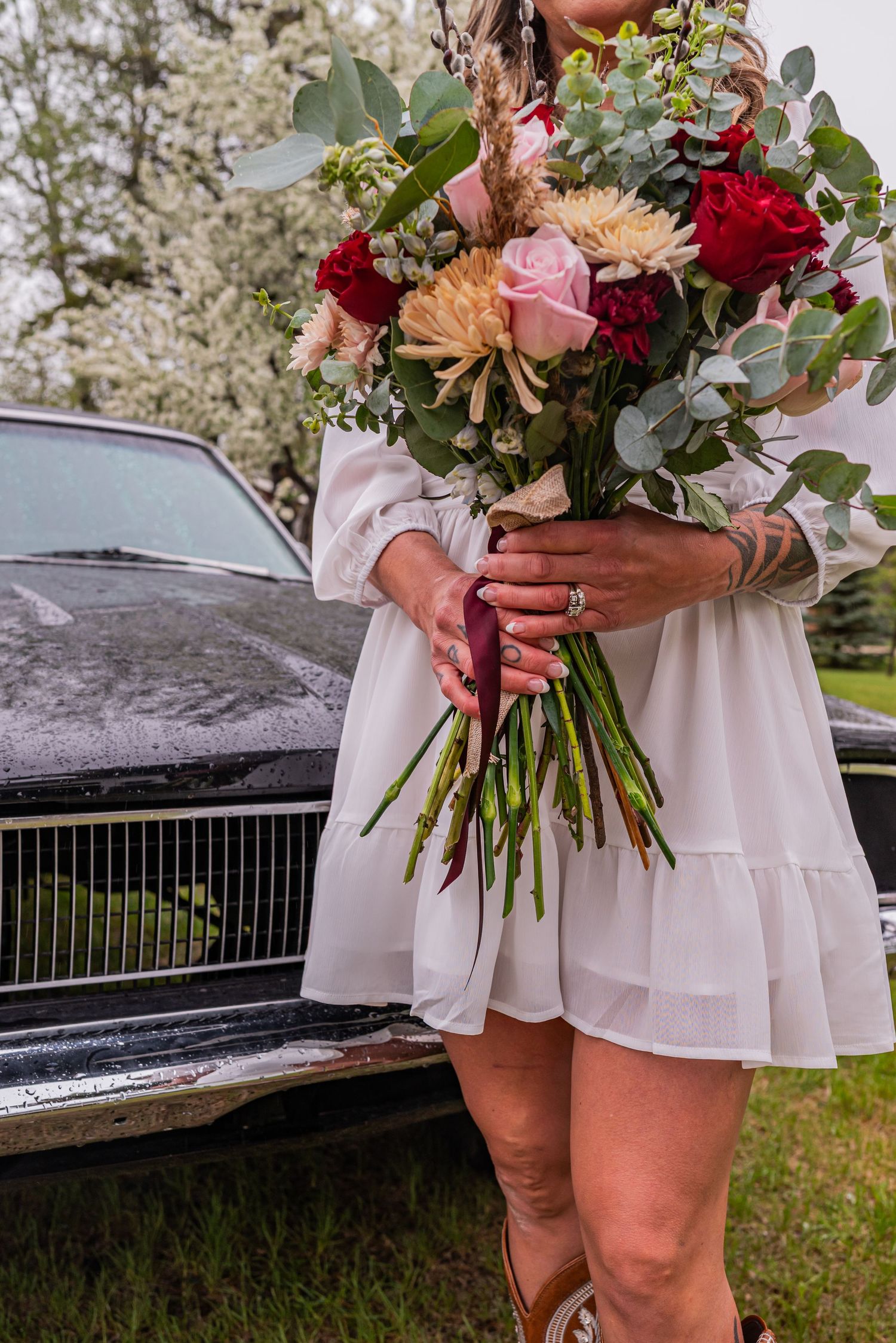 Woman in white dress holding a bridal bouquet of red roses and pink flowers beside a classic black car.