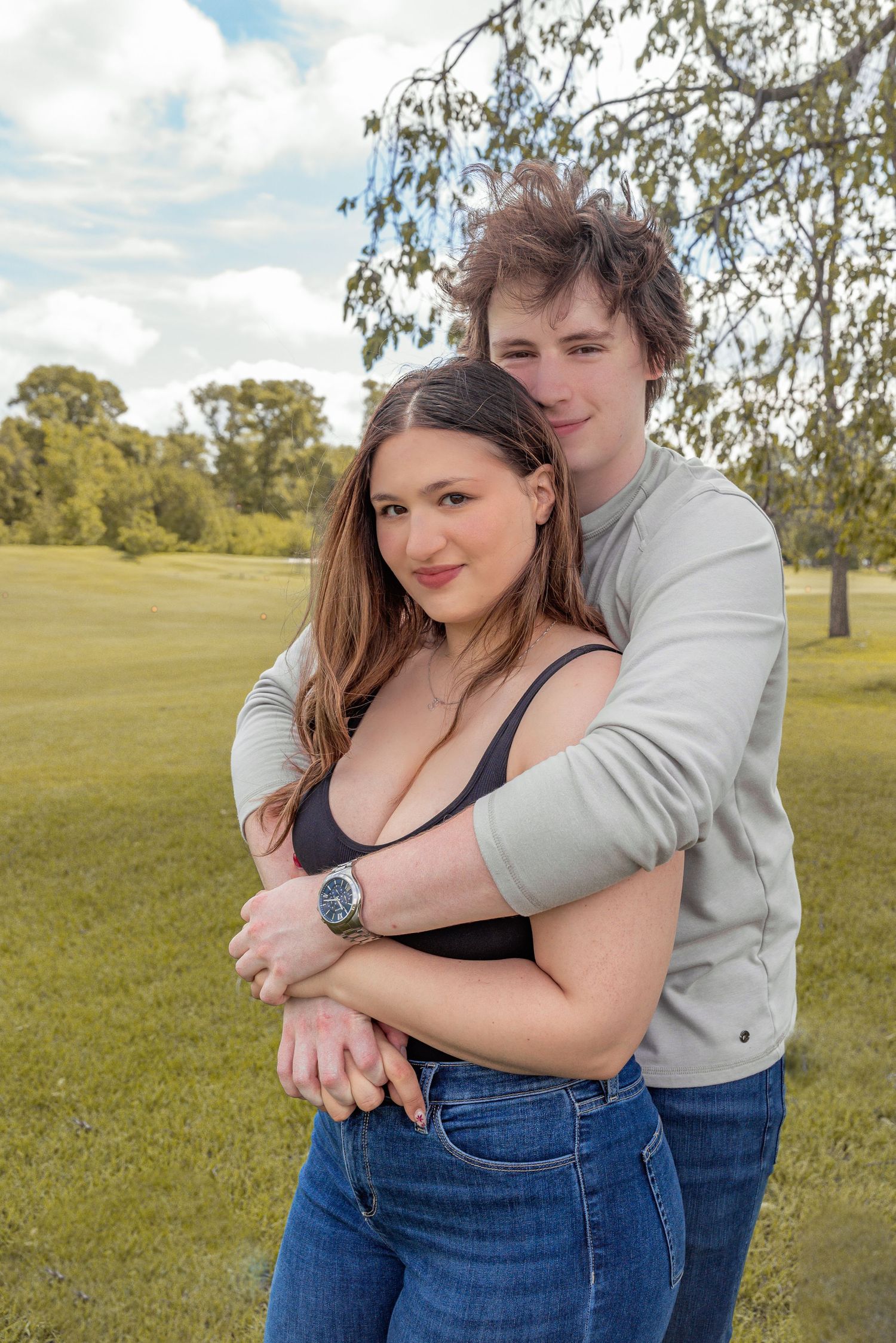 A young couple embracing outdoors in a green park, the man hugging the woman from behind, both wearing jeans.