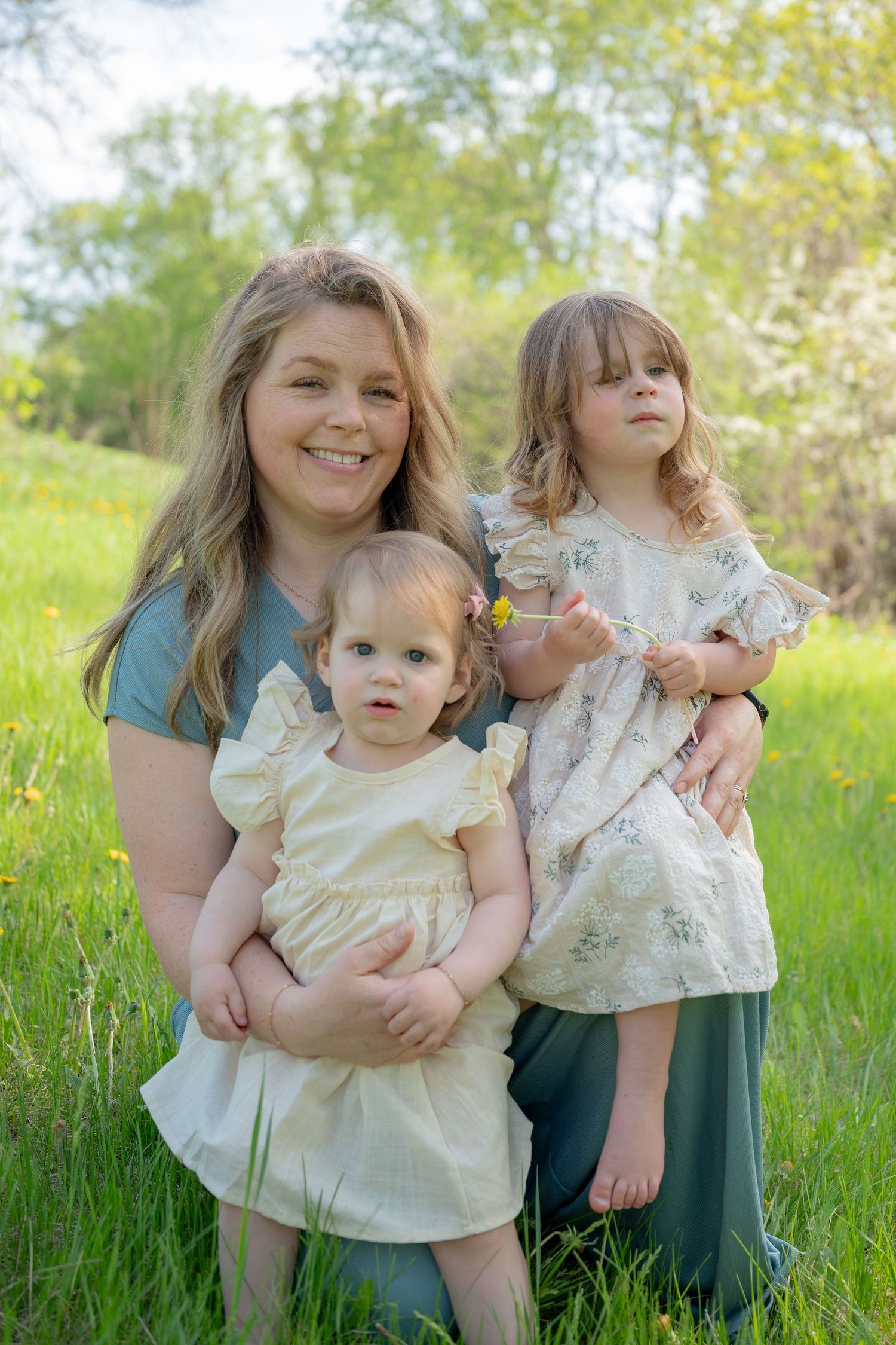 A smiling mother kneels in a green meadow with her two young daughters in floral dresses on a sunny spring day.