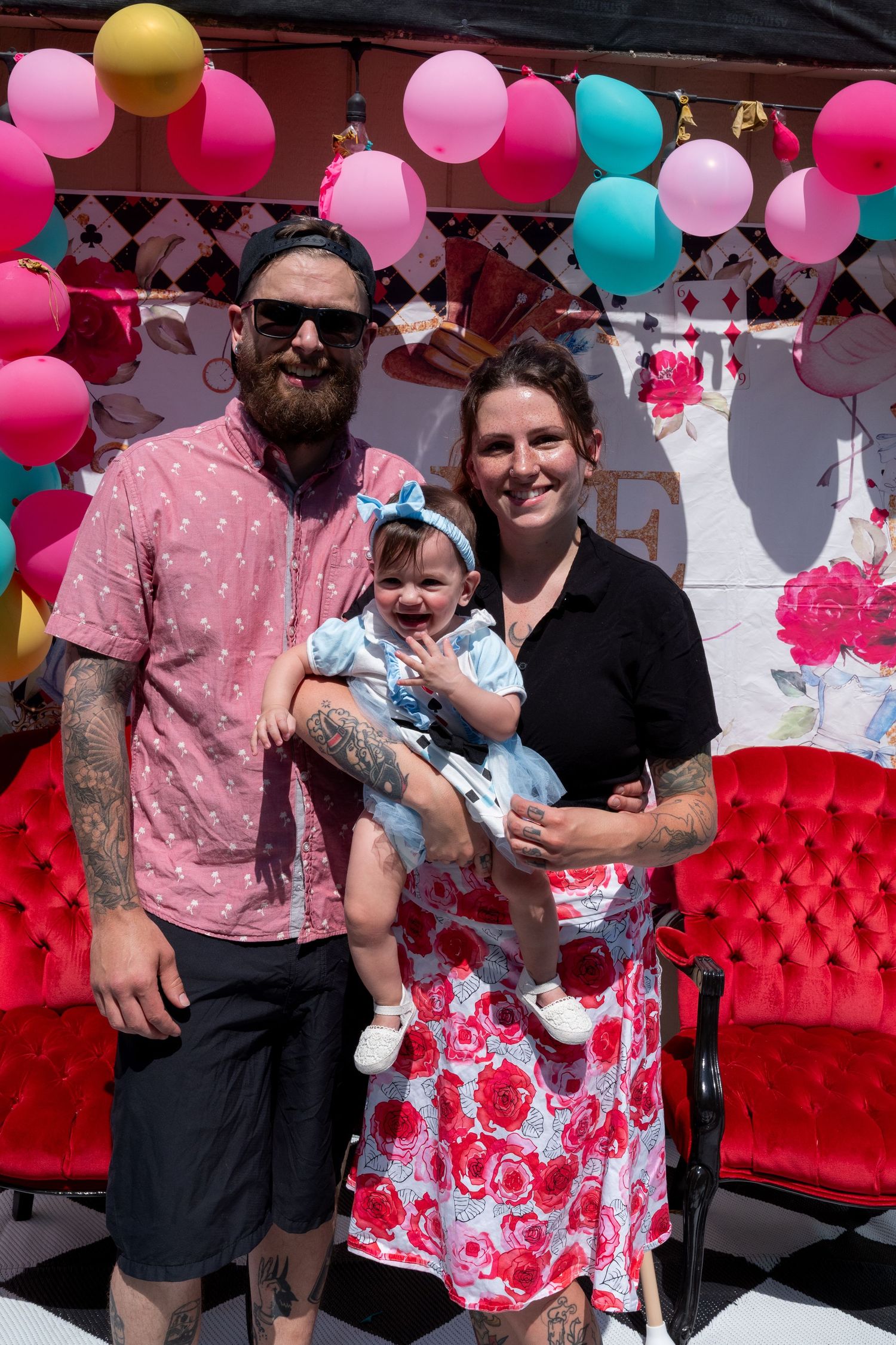 A couple with tattoos holds their smiling baby girl at a colorful balloon-decorated birthday party backdrop.