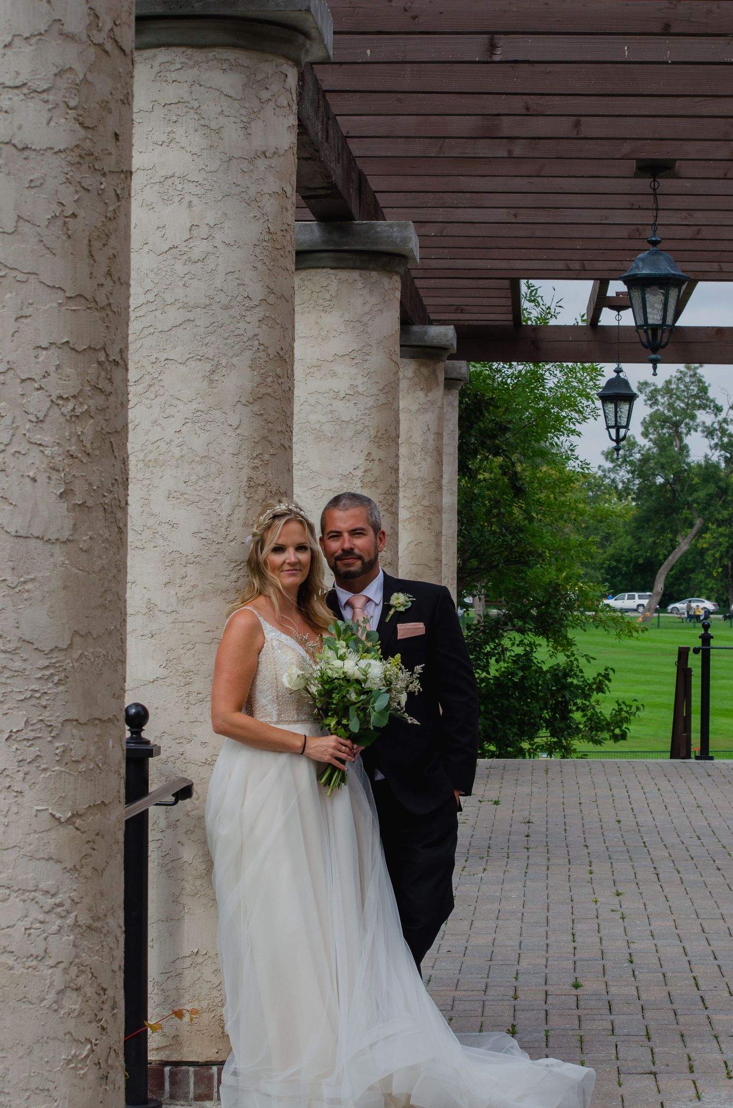 Bride in white gown holding bouquet stands with groom in black suit beside stone columns at elegant wedding venue.