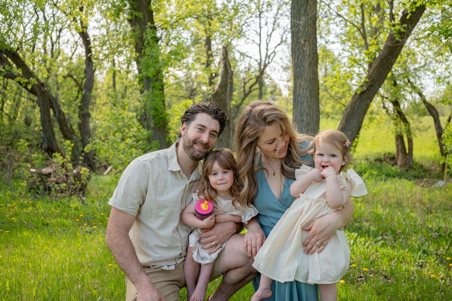 Happy family of four posing outdoors in lush green park, parents smiling with two young daughters on sunny day.