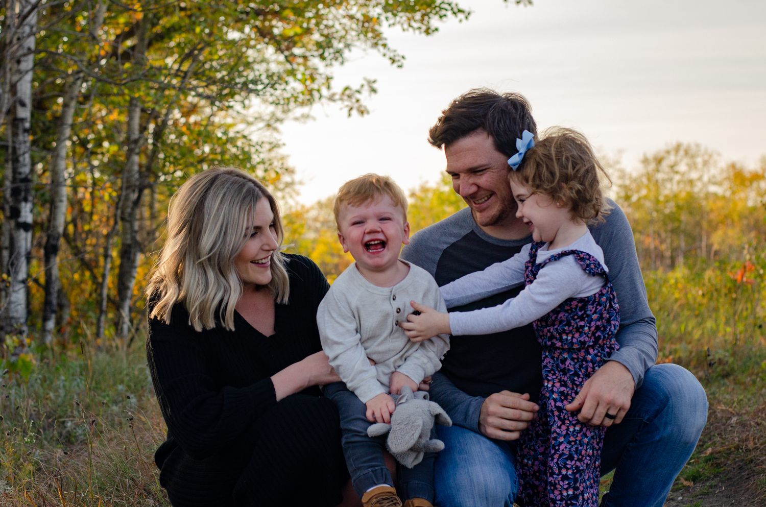 Family of four smiling outdoors in autumn, parents holding toddlers among golden trees at sunset.