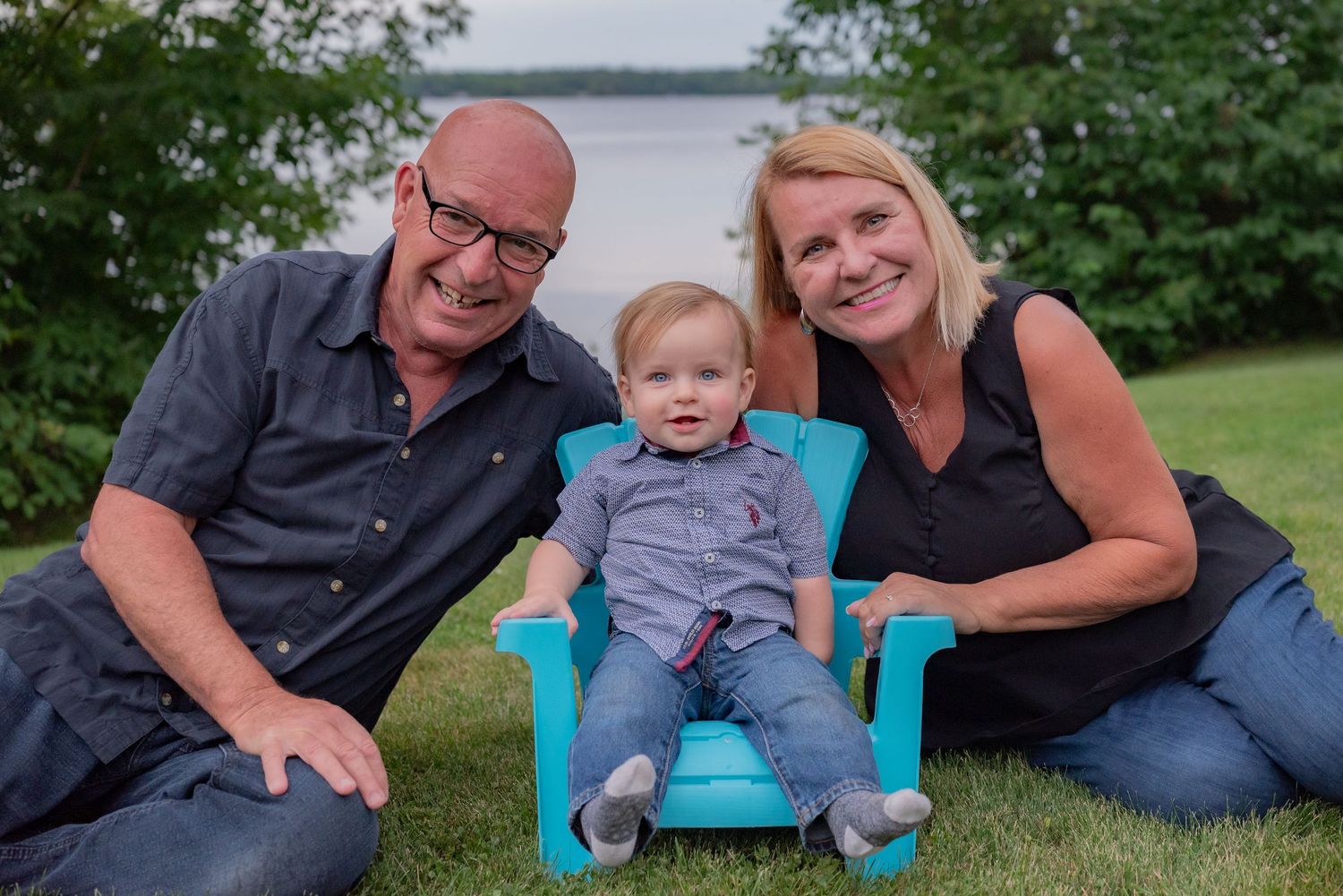 Family portrait of two smiling adults sitting on grass with a baby in a teal chair, near a lake.