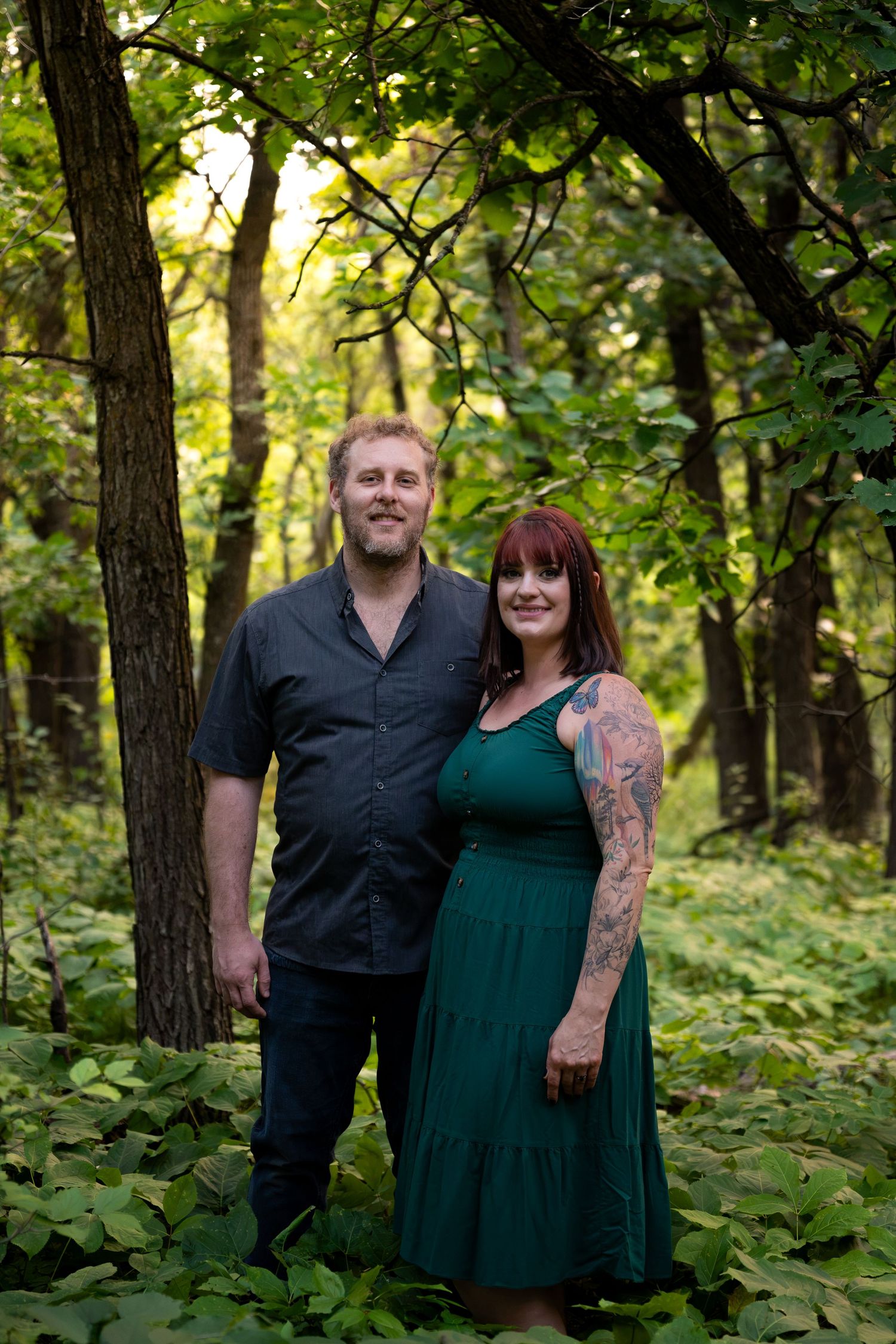 A couple smiles together in a lush green forest, woman in teal dress with tattoos, man in dark shirt.
