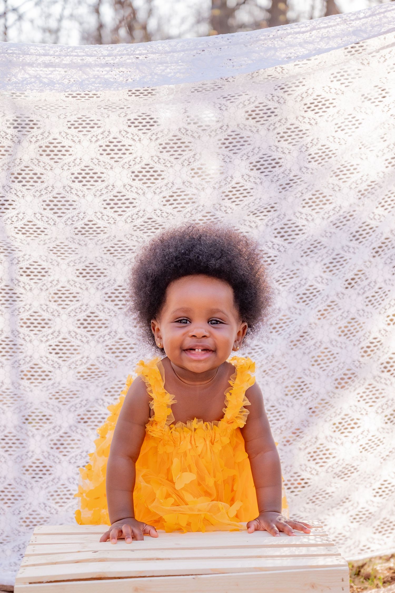 Smiling Black toddler girl in yellow dress with afro hair standing against white lace backdrop outdoors.
