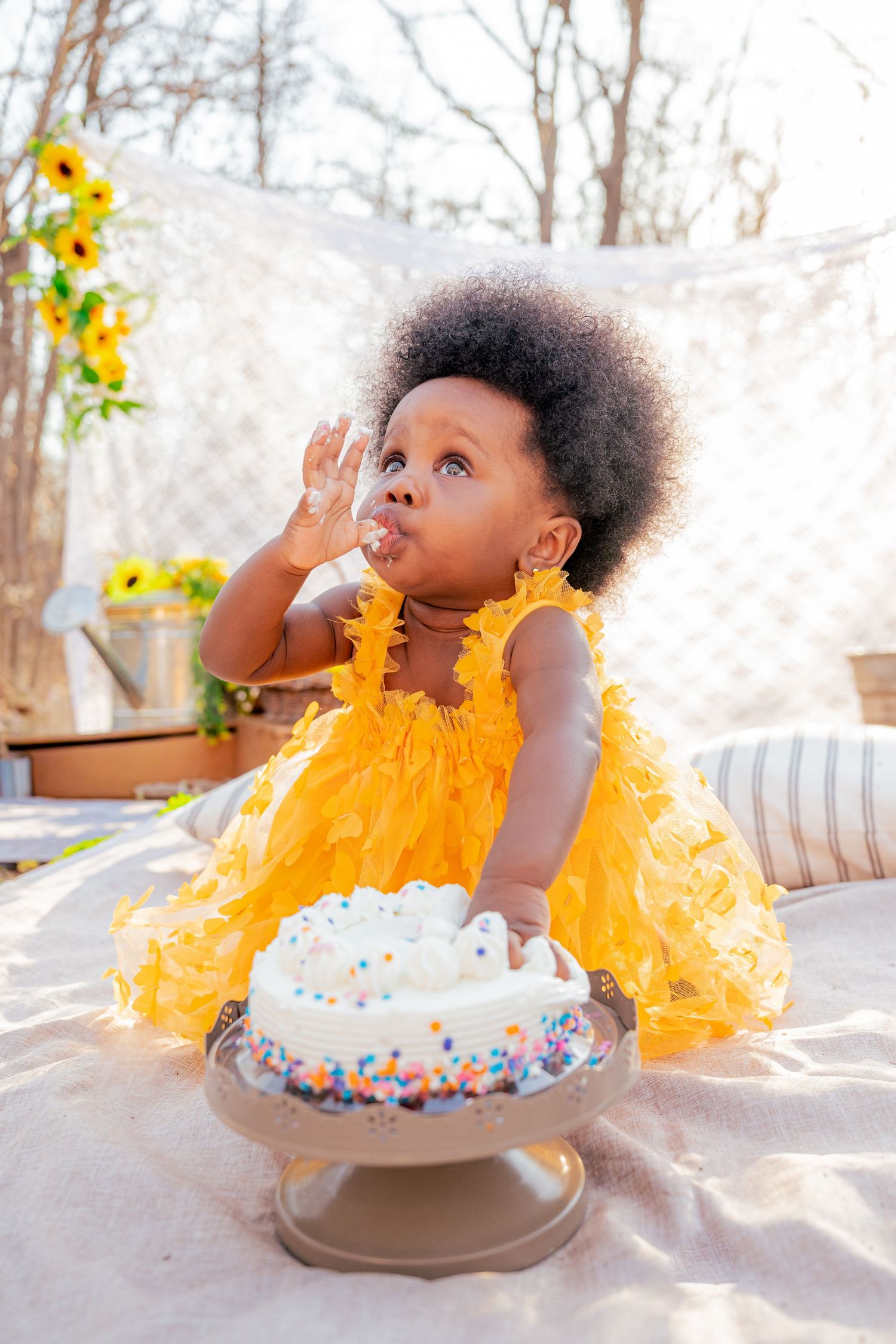 Adorable baby girl in yellow ruffled dress sitting with white sprinkle birthday cake during first birthday cake smash photoshoot.