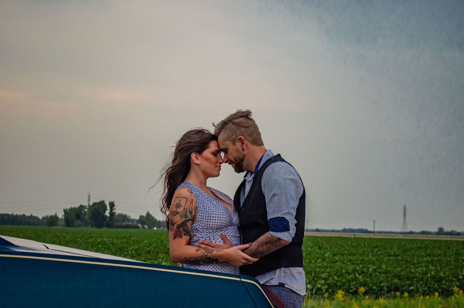 A couple shares a romantic moment near a vintage car with green fields in the background on a cloudy day.