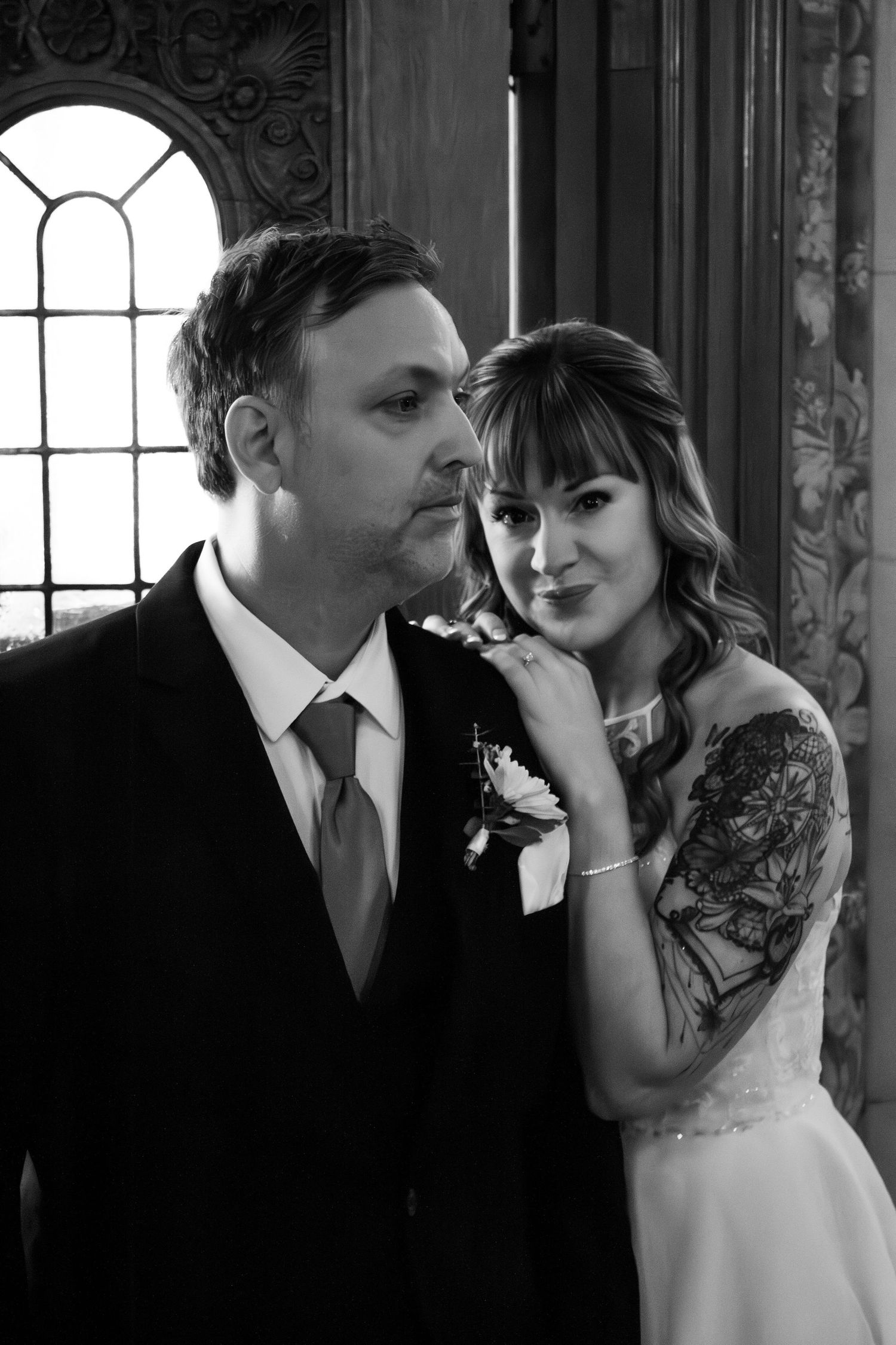 Black and white wedding portrait of bride with tattoo and groom in suit with boutonniere posing by ornate window.