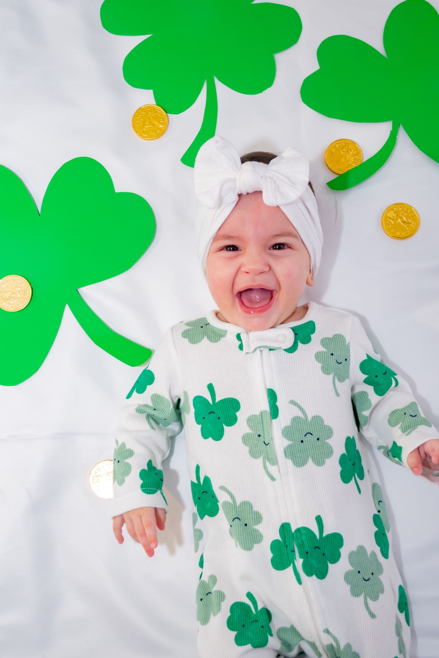 Happy baby wearing shamrock print onesie and white bow headband surrounded by green clover cutouts and gold coins for St. Patricks Day.