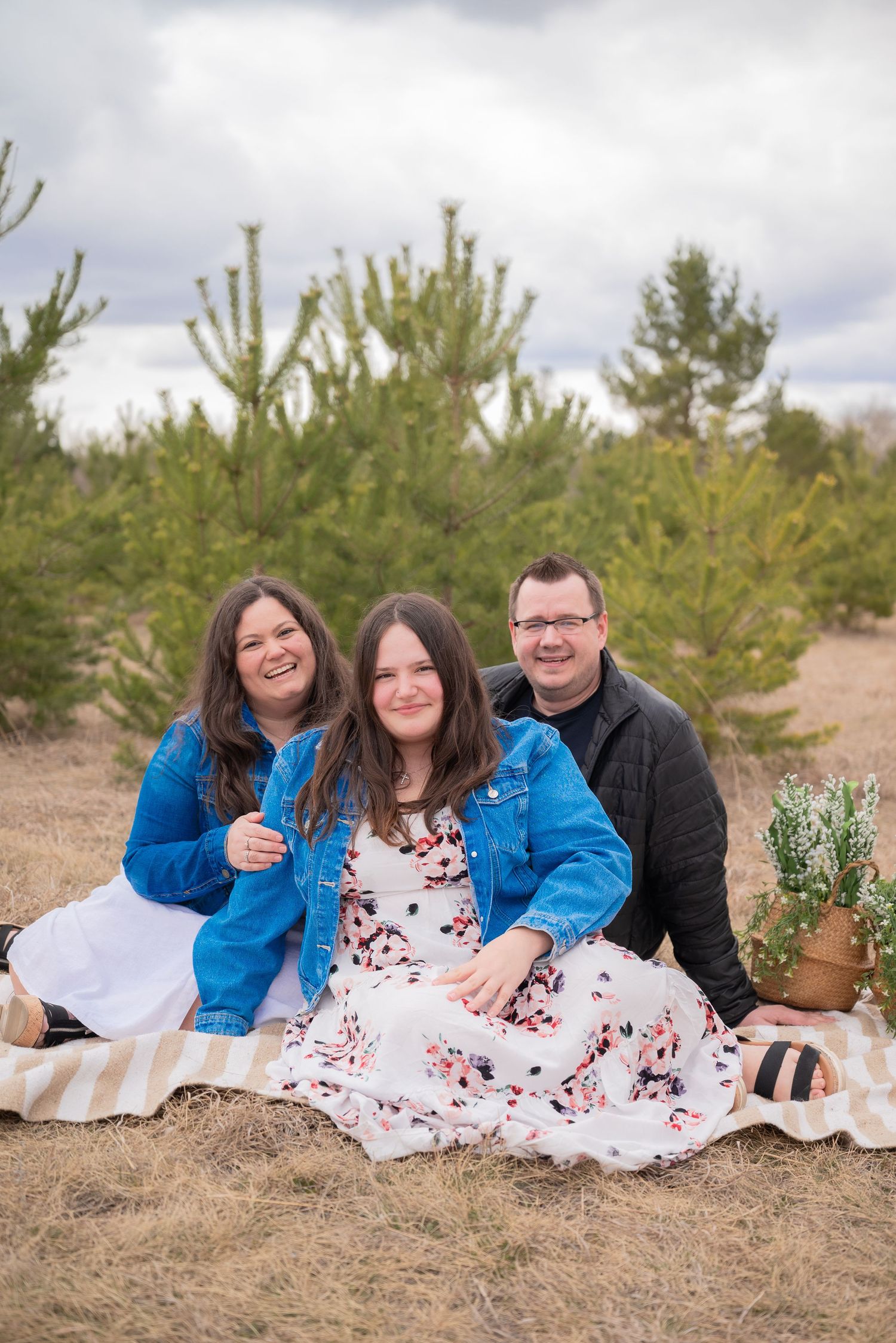 Family of three smiling together on a blanket outdoors near pine trees, wearing denim jackets in a casual portrait.
