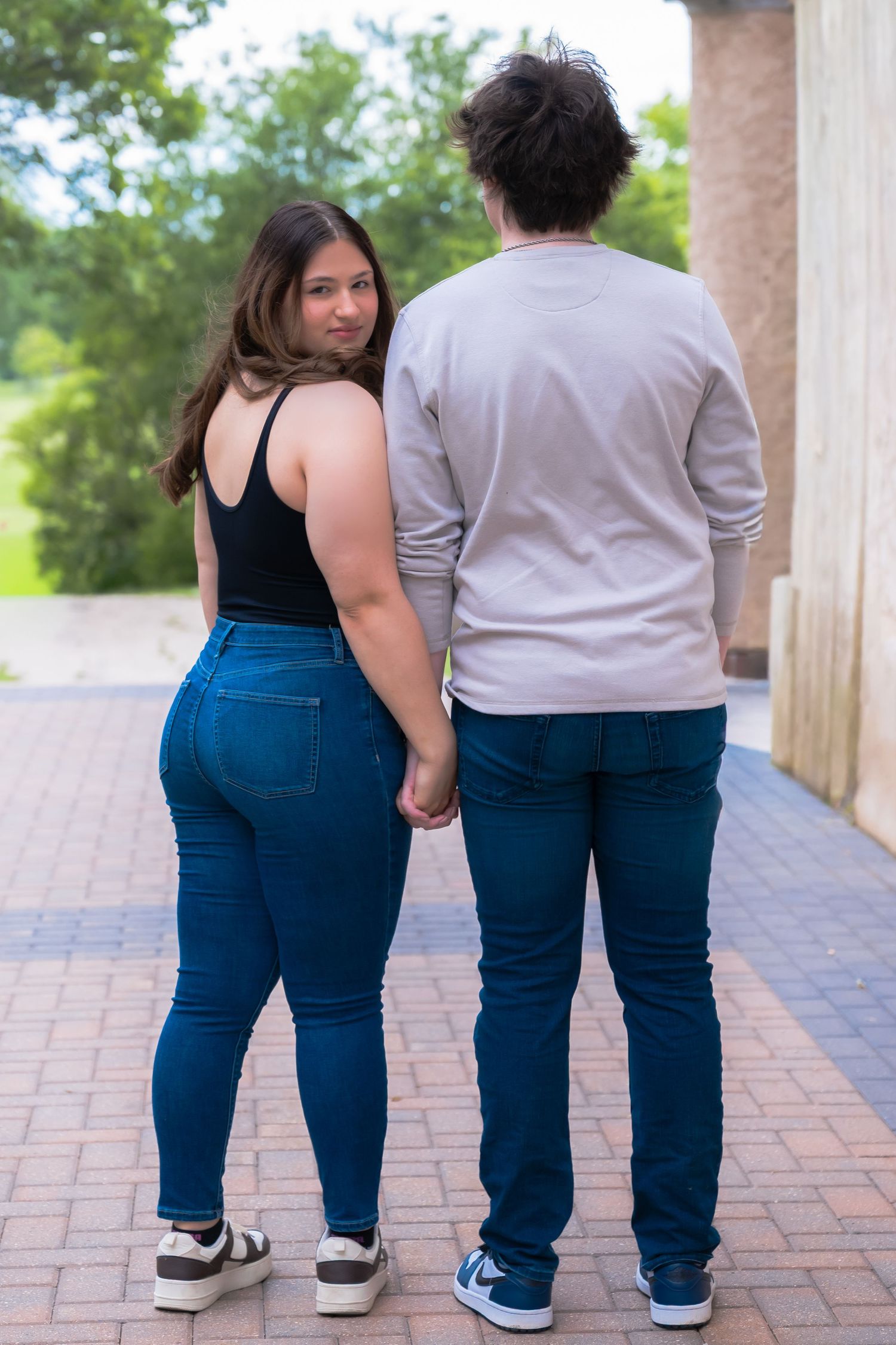Couple holding hands walking away on brick path, woman in black top and jeans looks back smiling.