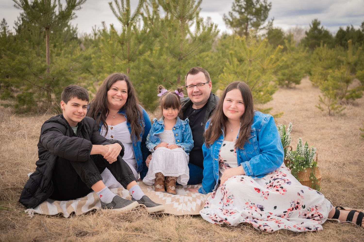 Family of five smiling together outdoors in a pine forest setting, sitting on dry grass for a portrait.