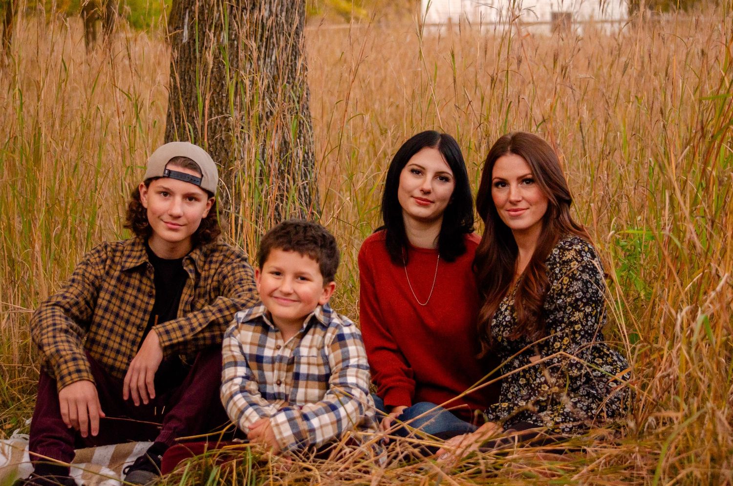 Family of four smiling together in a golden autumn field, sitting near a tree for a warm fall portrait.