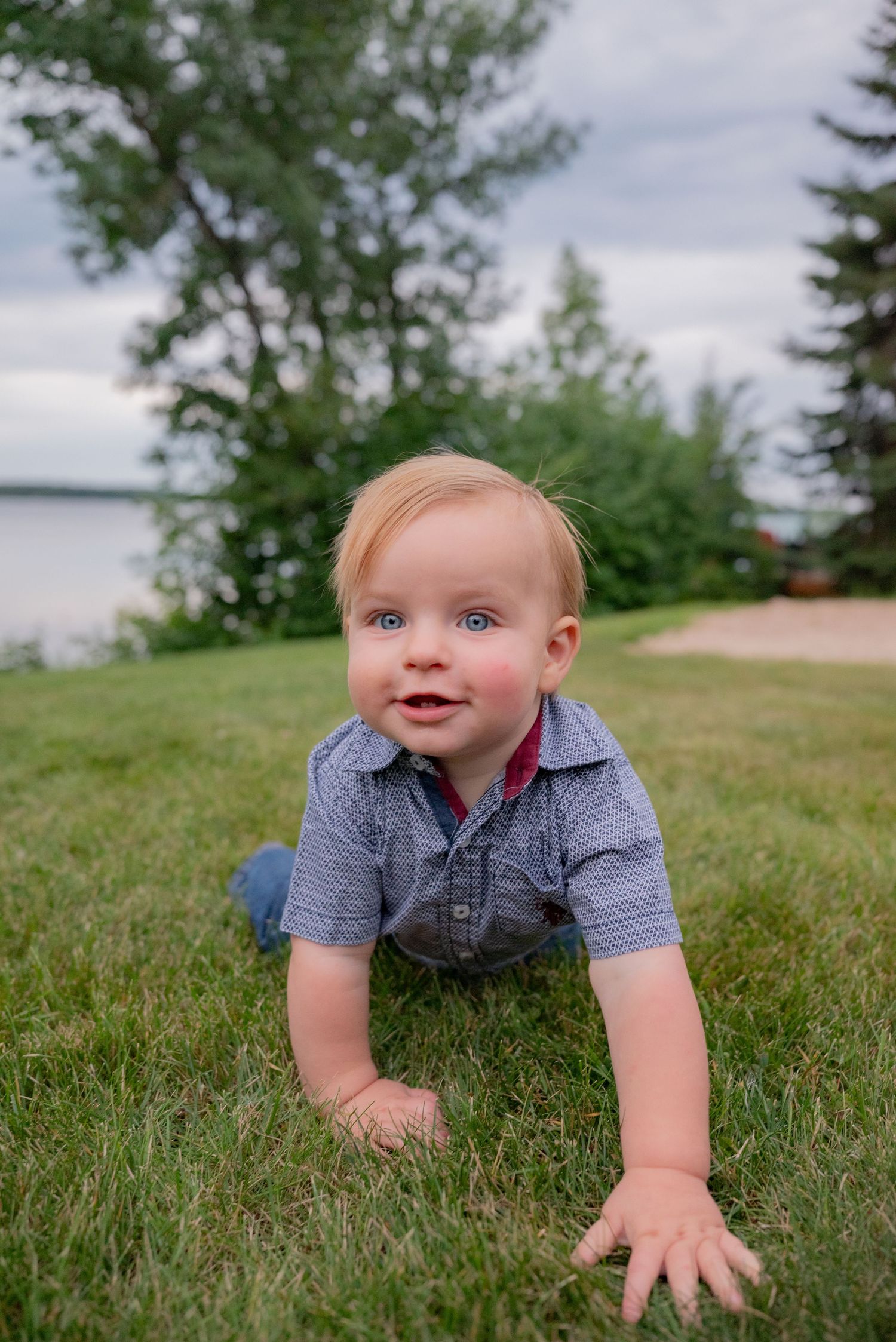 A blue-eyed baby boy crawls on green grass outdoors, wearing a gray button-up shirt, smiling at the camera.