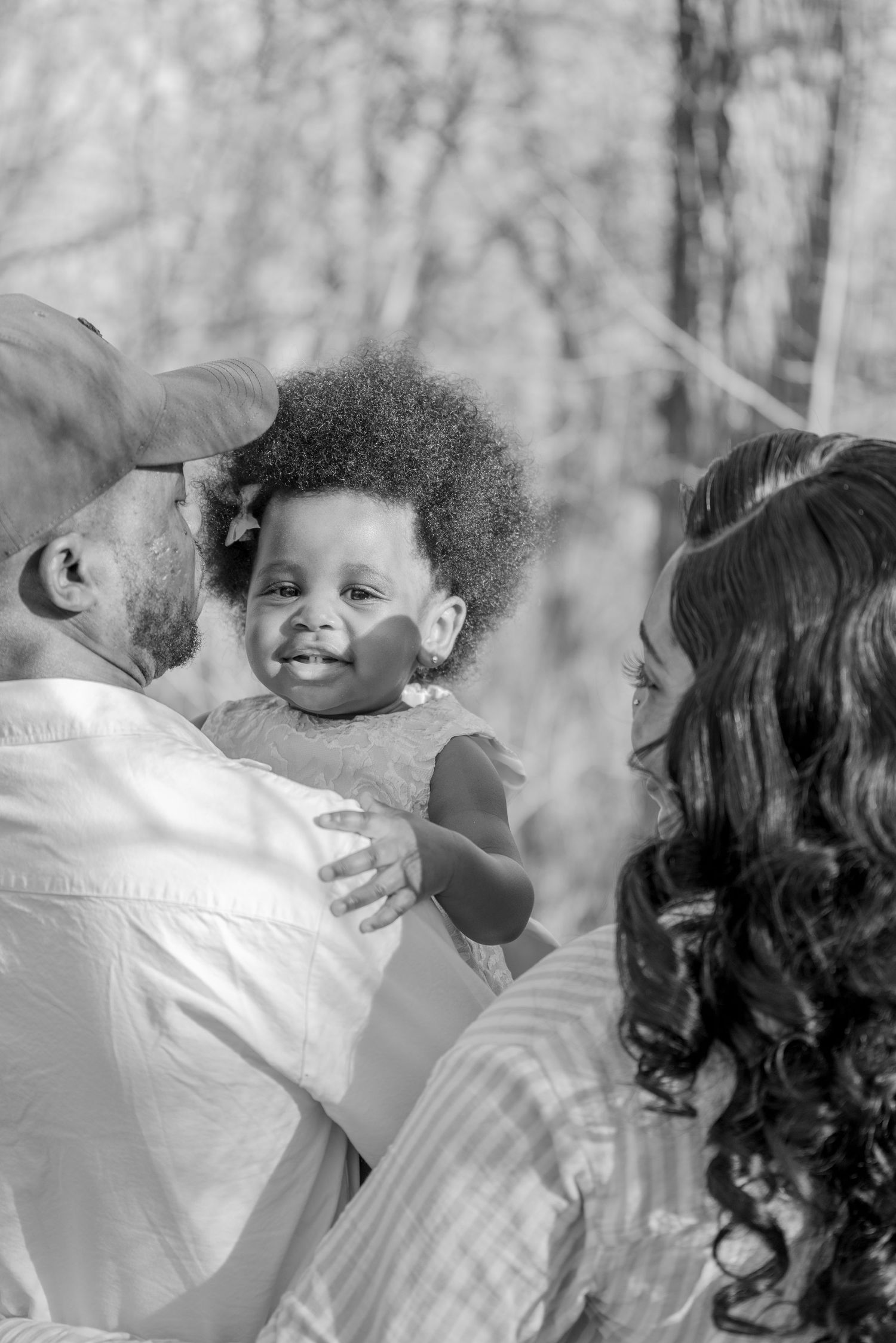 Black and white photo of parents lovingly holding their smiling baby girl outdoors among trees.