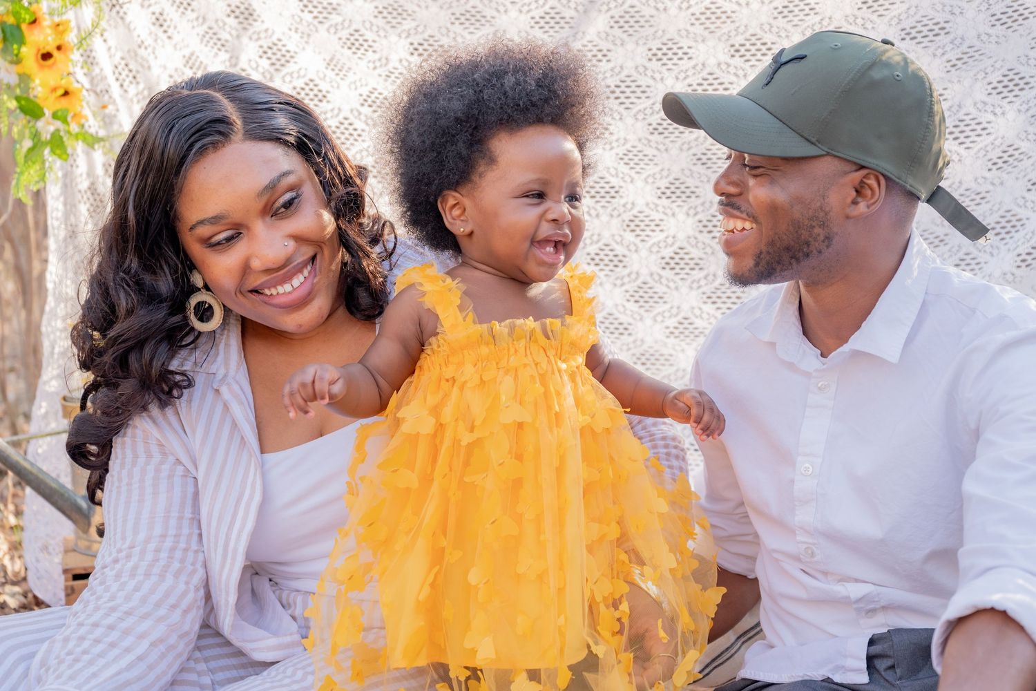 A happy Black family with a smiling mother, joyful toddler in yellow dress, and father in green cap posing together.