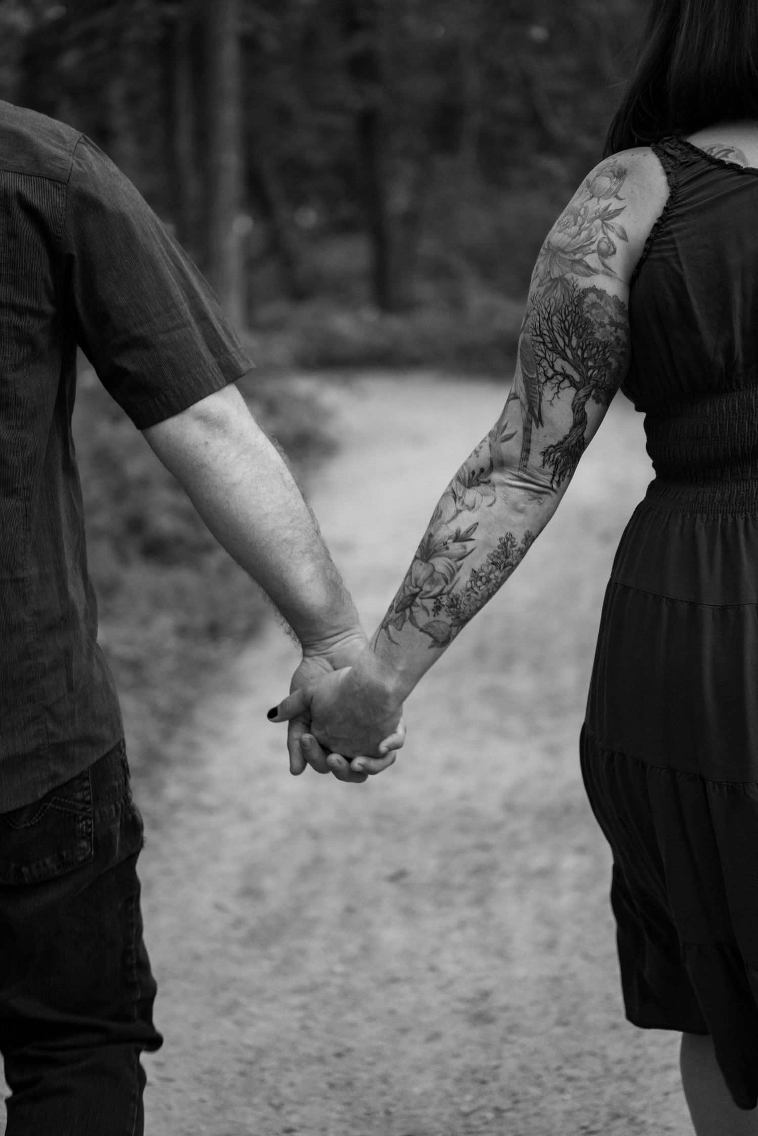 Black and white photo of a couple holding hands on a forest path, woman with floral tattoo sleeve.