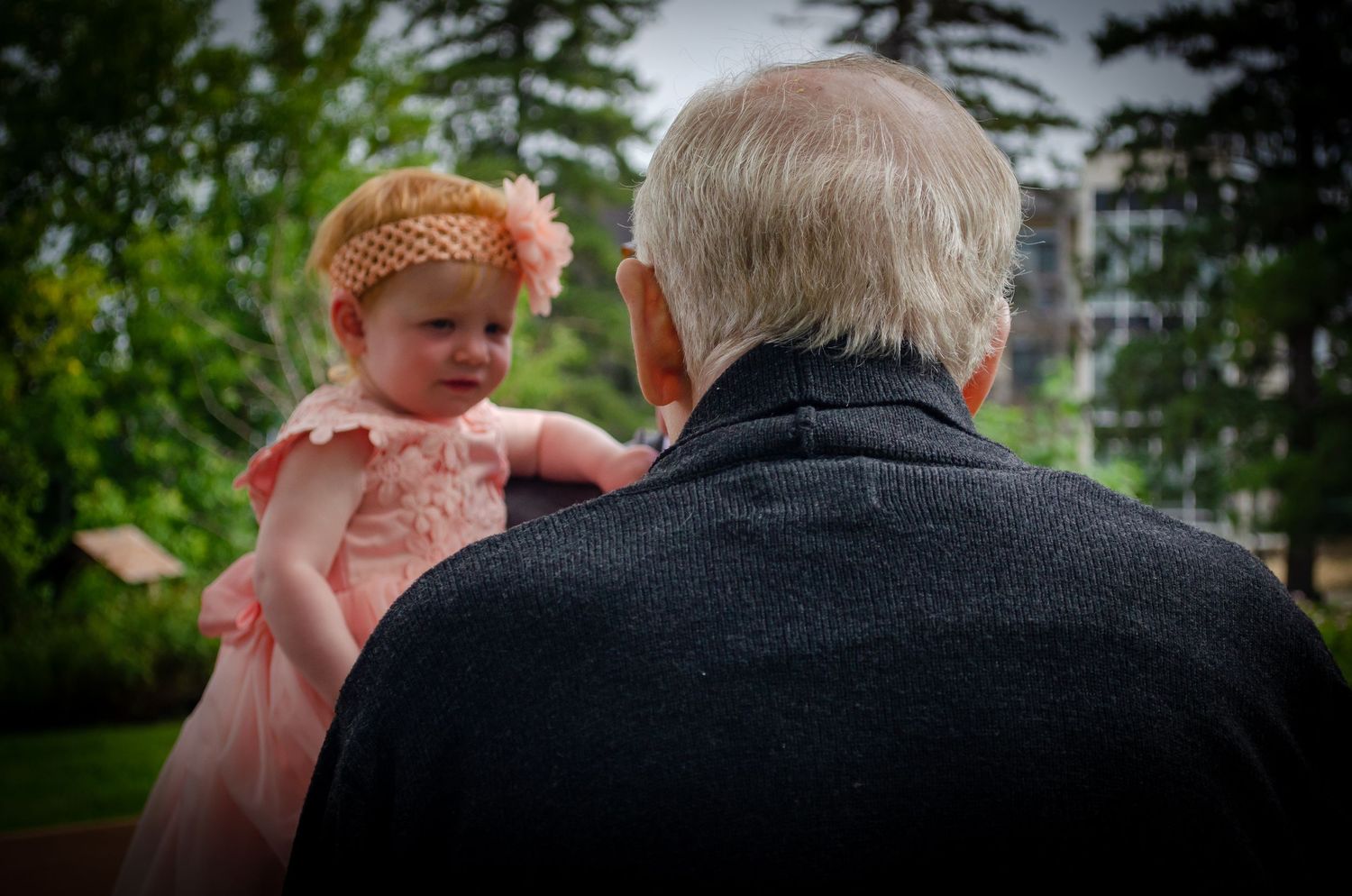 Elderly man holding a baby girl in a pink dress and headband outdoors in a lush green garden setting.