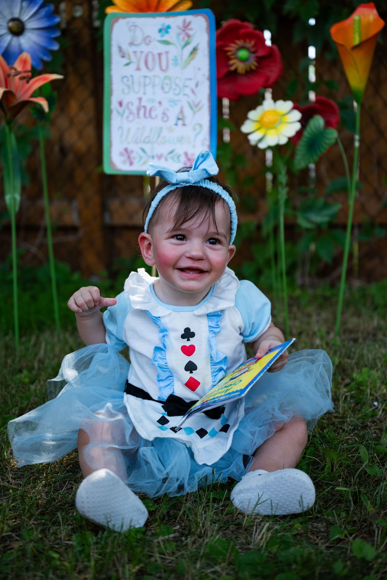 Baby dressed as Alice in Wonderland sits on grass with giant flowers and a Mad Hatter sign backdrop.