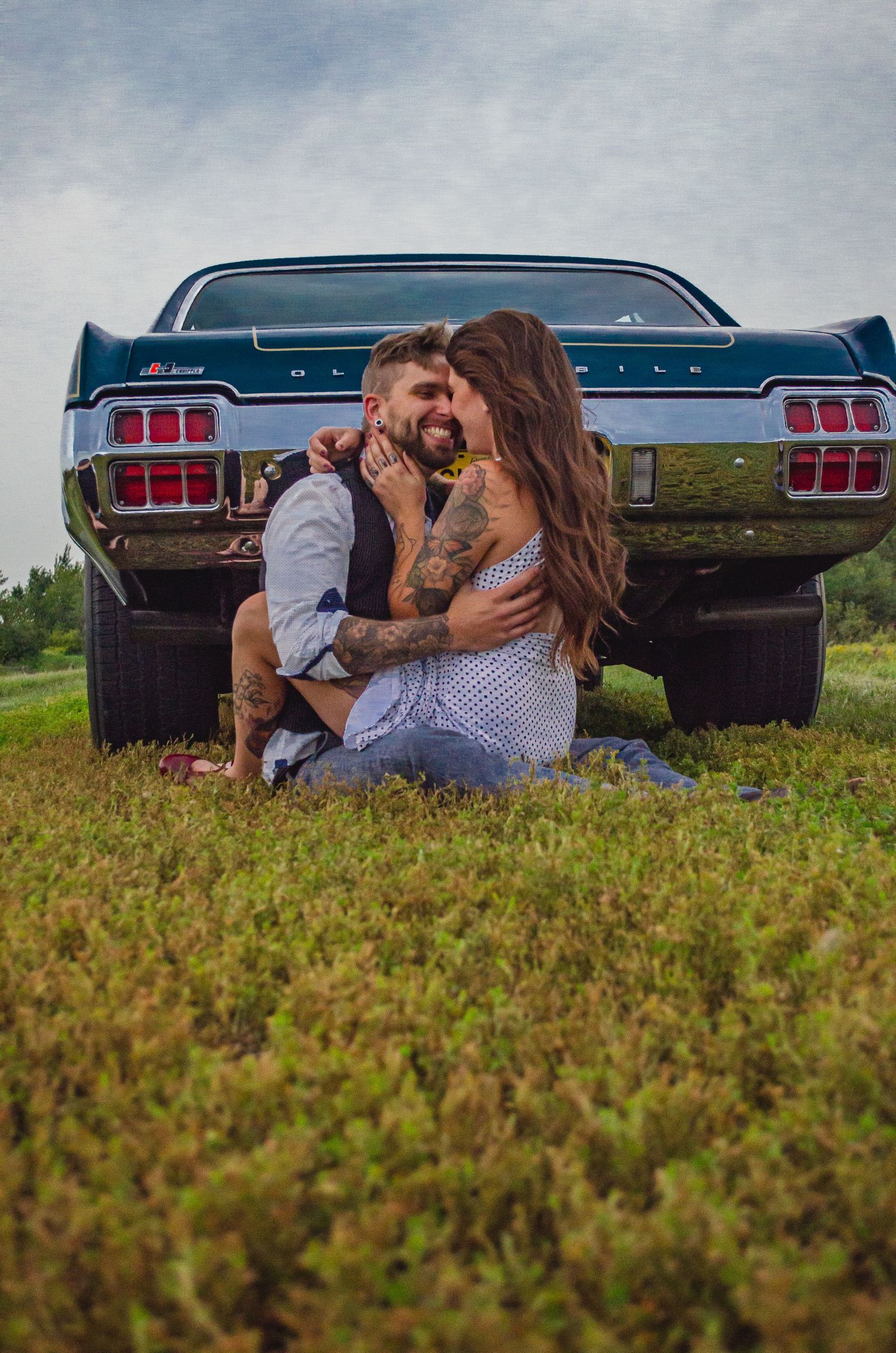 Tattooed couple sharing a romantic kiss sitting in grass behind a classic vintage teal muscle car in an open field.