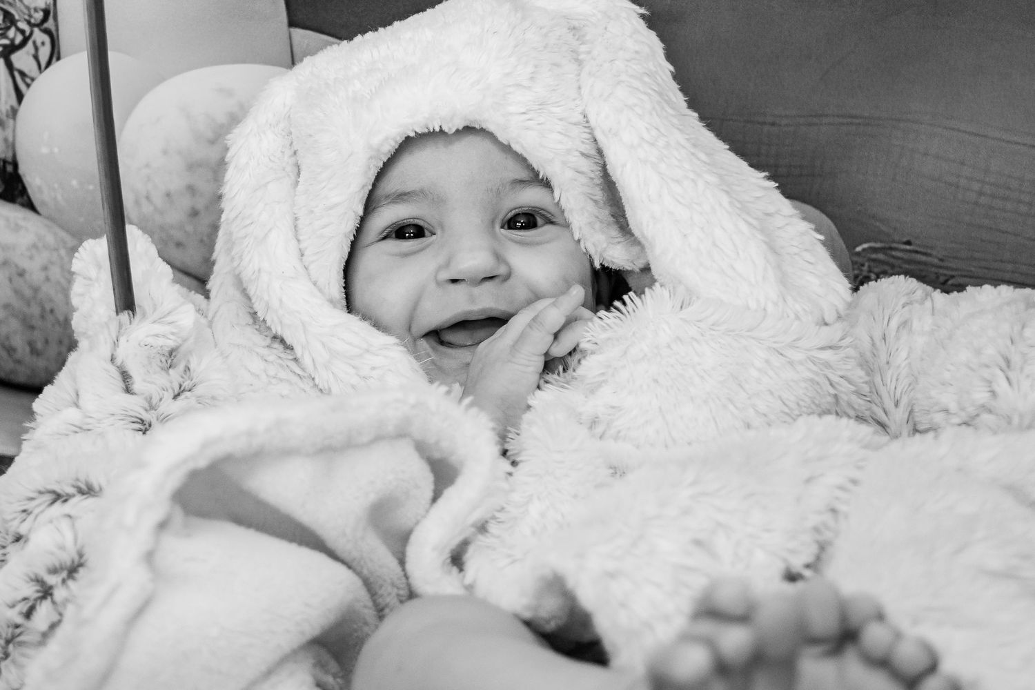 Black and white photo of a smiling baby wrapped in a fluffy white bunny hooded blanket, lying on a soft surface.