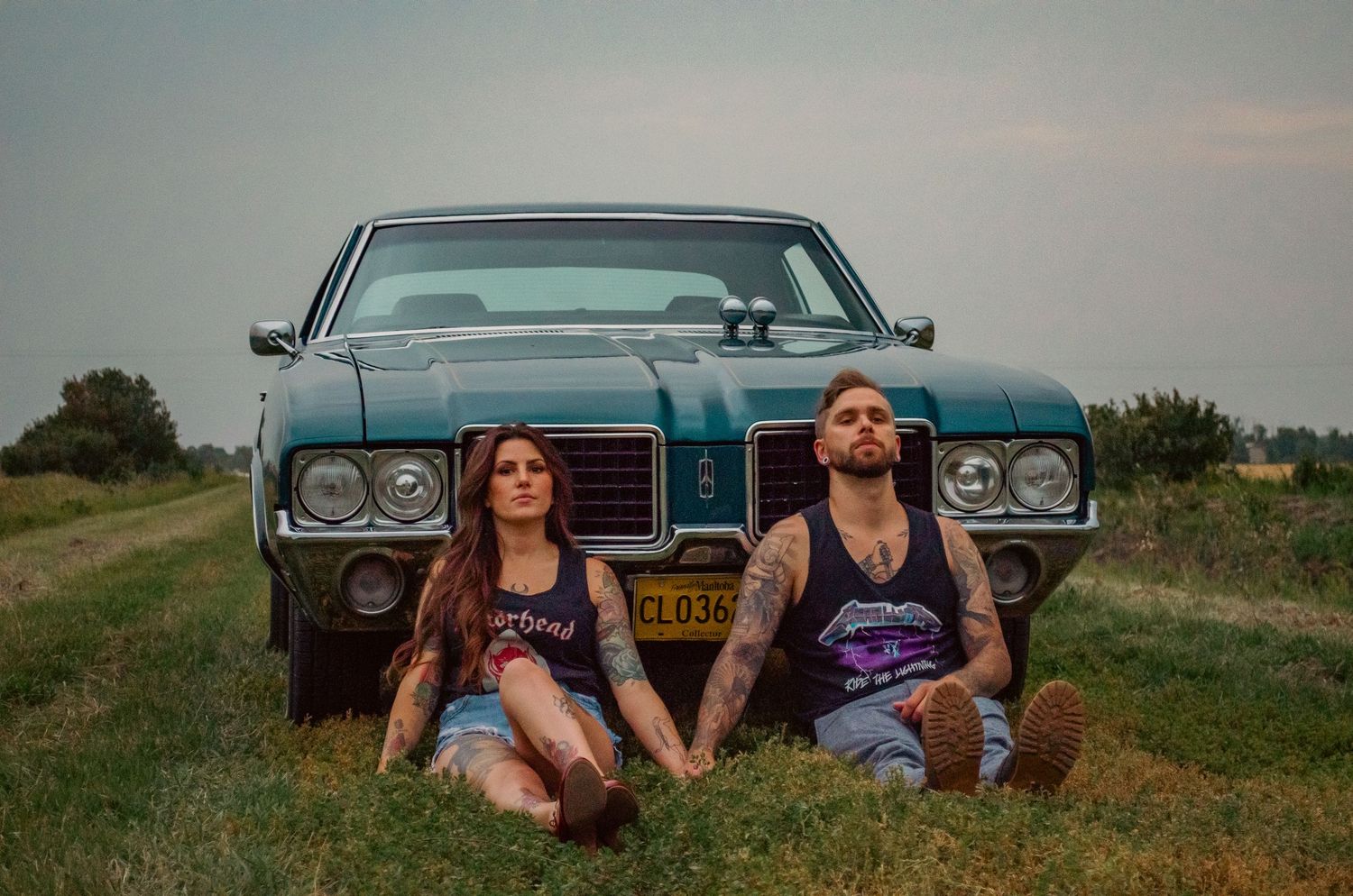 A man and woman sit in front of a vintage teal muscle car on a grassy field, both wearing rock band t-shirts.