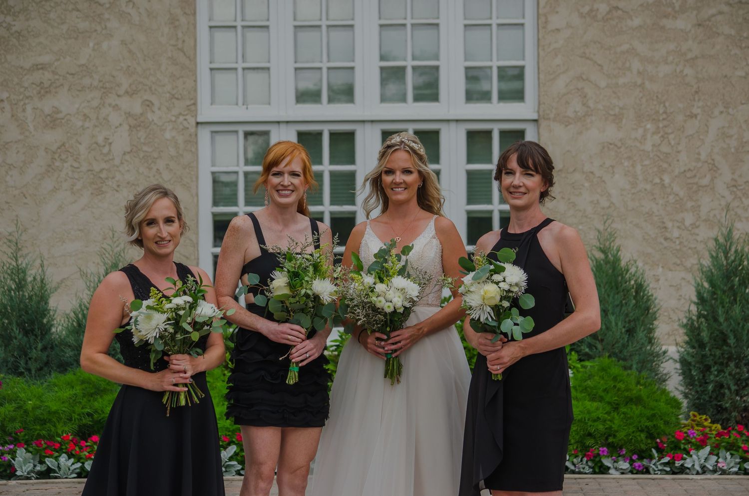 Bride in white gown with three bridesmaids in black dresses holding green and white bouquets outside elegant venue.