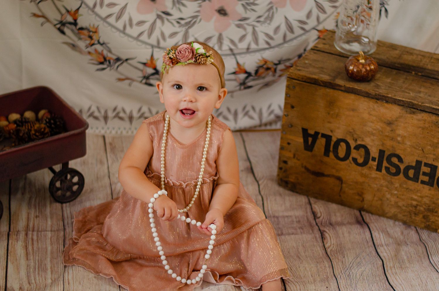 Baby girl in pink dress with pearl necklace sits beside vintage Pepsi-Cola crate on rustic wood floor.
