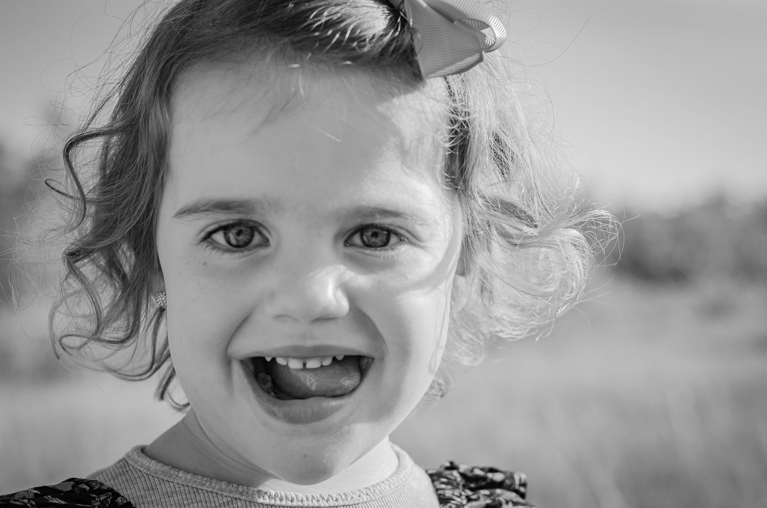Black and white portrait of a smiling toddler girl with curly hair and a bow outdoors.