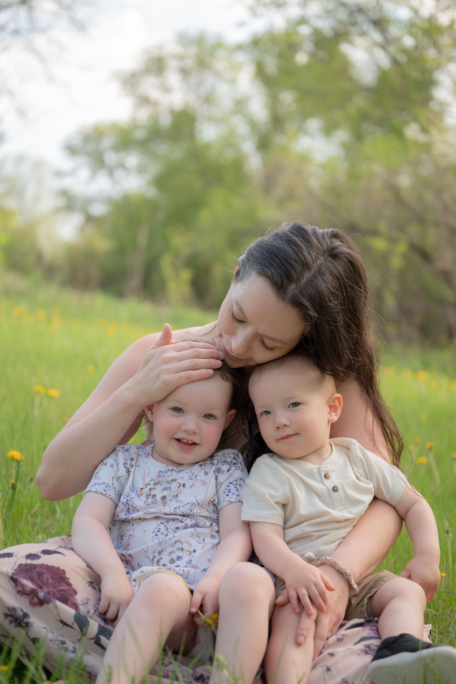 A mother sits in a sunny green field, tenderly kissing her two smiling toddlers on a warm spring day.