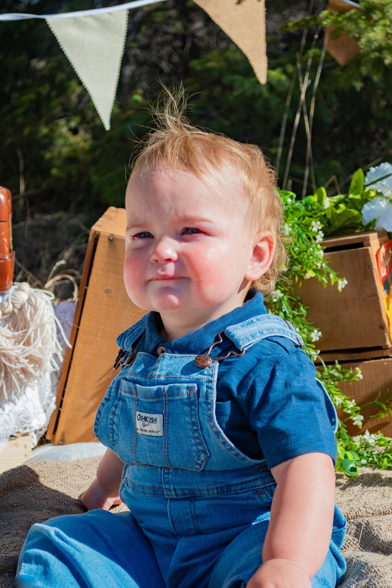 A cute baby in denim overalls sits outdoors making a funny face at a rustic birthday party setup.