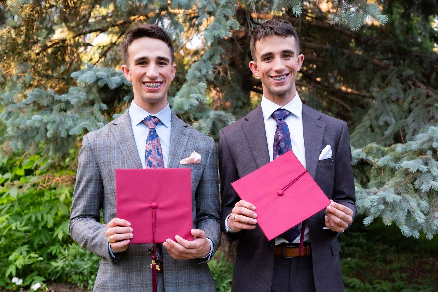 Two young men in suits holding red graduation folders smile outdoors among green trees.