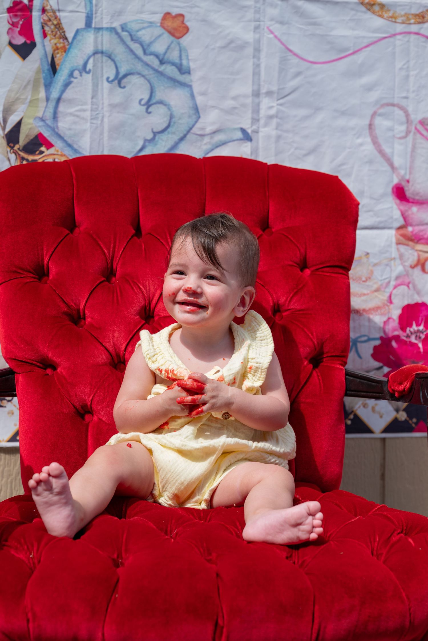 Happy baby girl in yellow dress sitting on a red tufted velvet chair, smiling with messy hands.