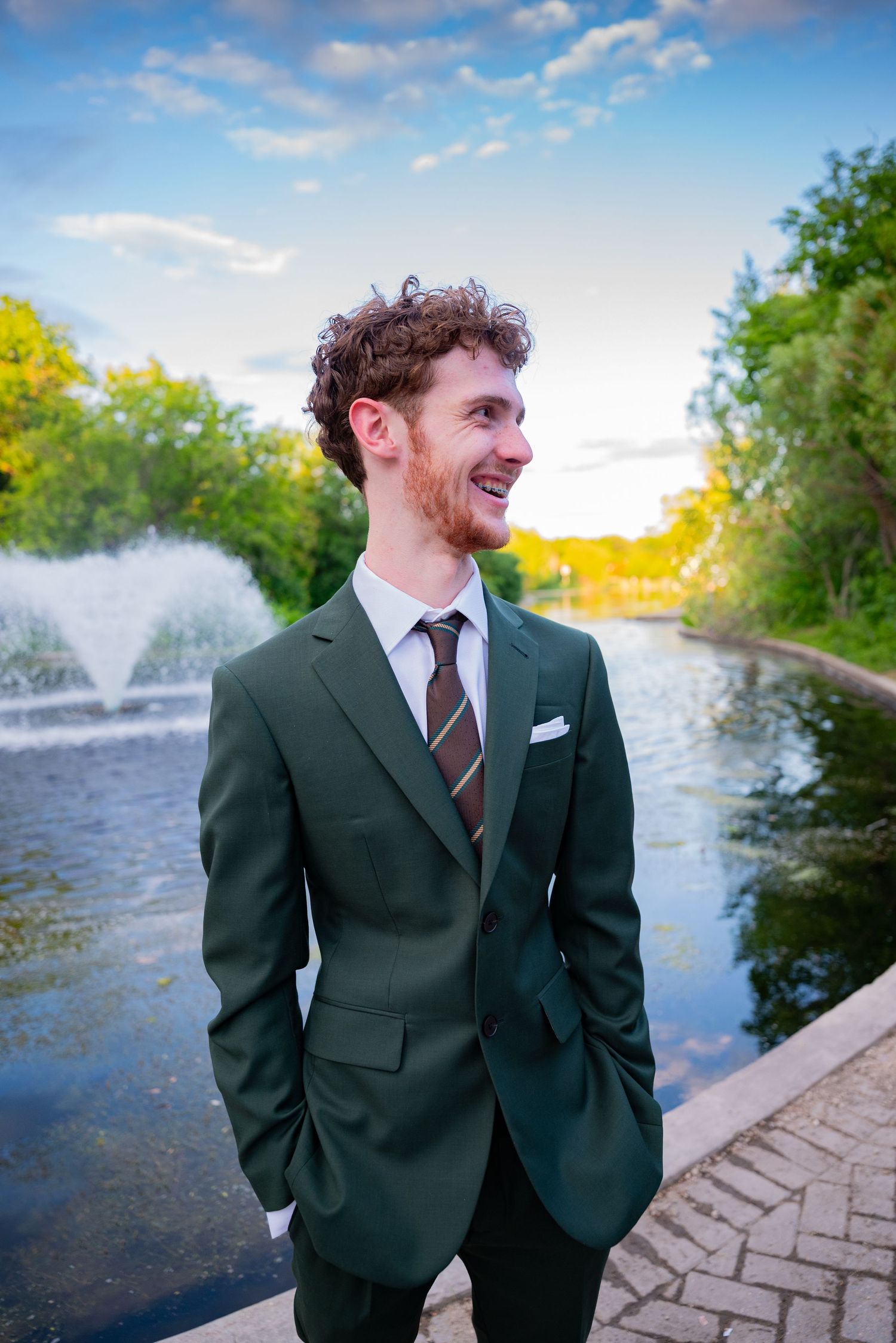 Young man in green suit and striped tie smiling outdoors near a fountain and pond.