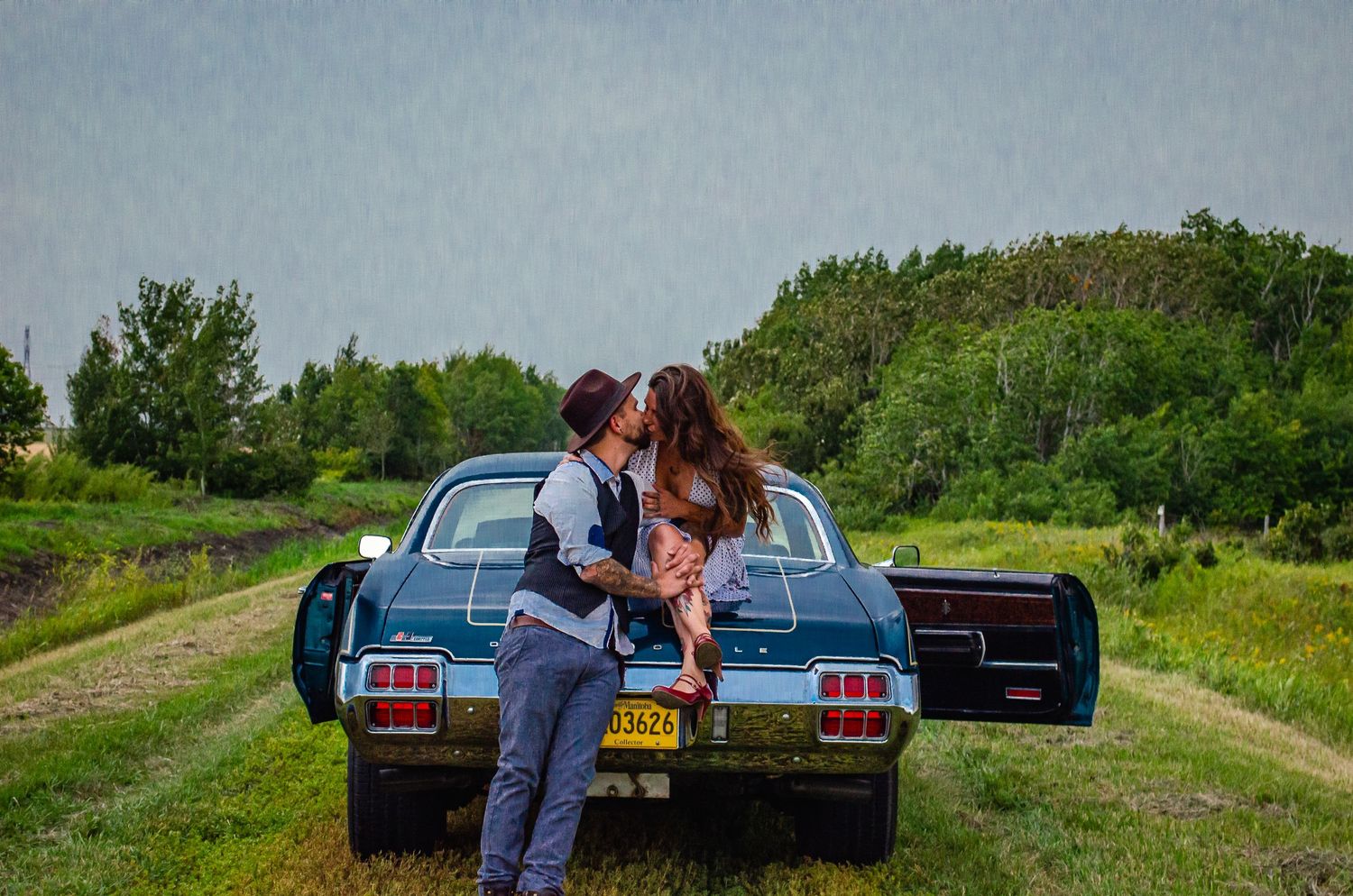 Couple kissing beside a vintage teal car on a rural dirt path surrounded by lush green trees and grass.