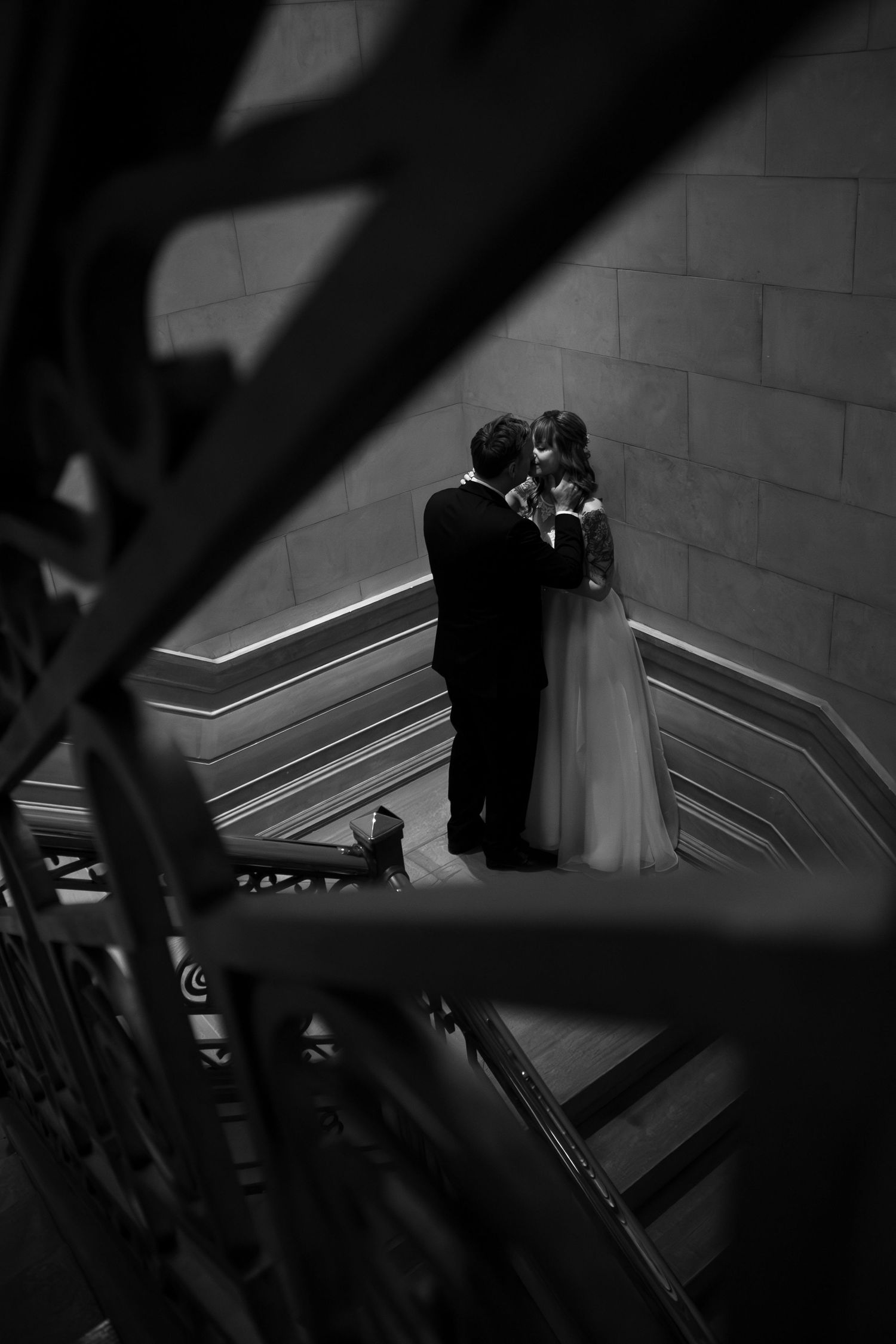 Black and white wedding photo of bride and groom embracing on grand staircase, viewed through ornate iron railings.