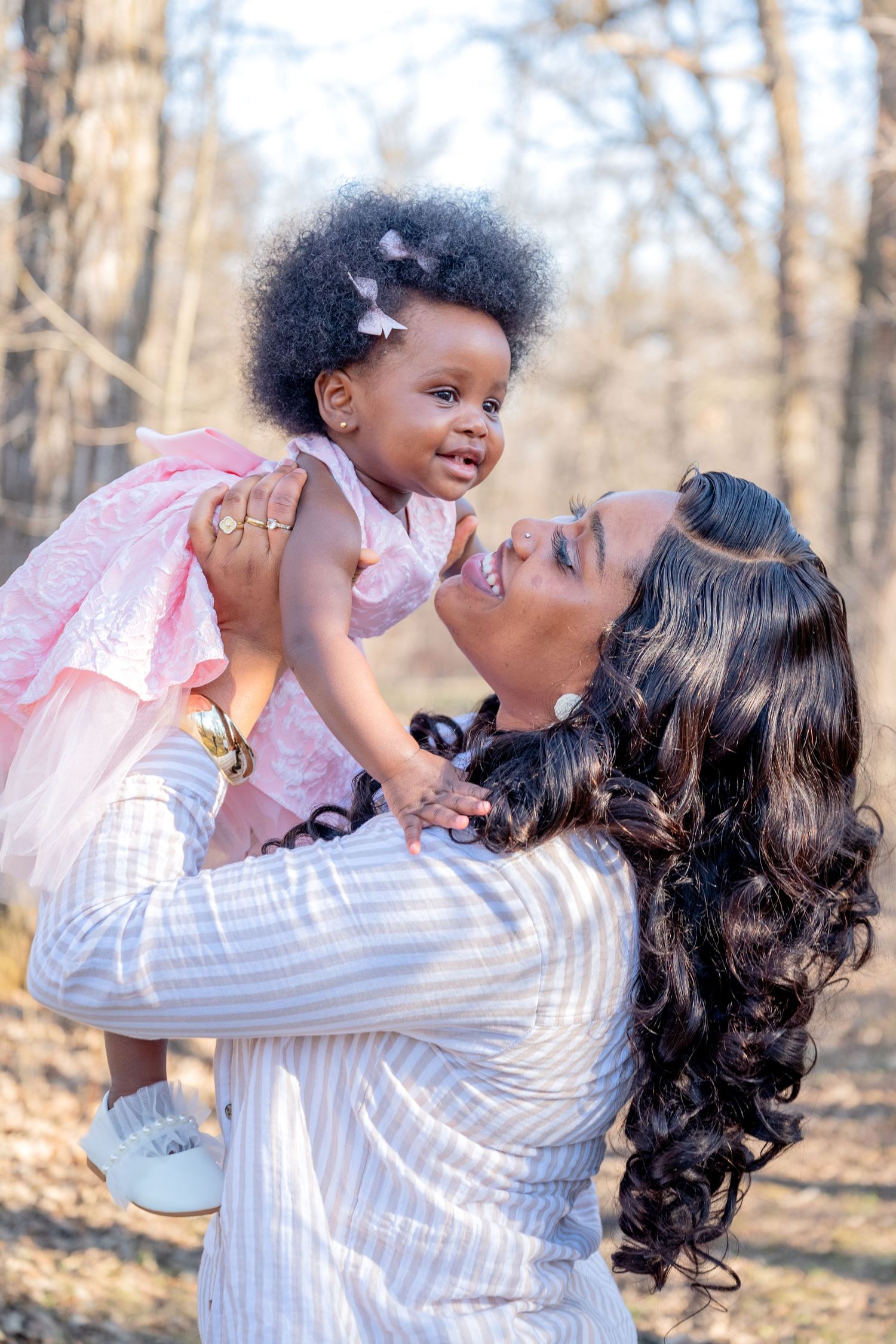 A mother lovingly holds her baby girl in a pink dress outdoors among bare trees in soft natural light.