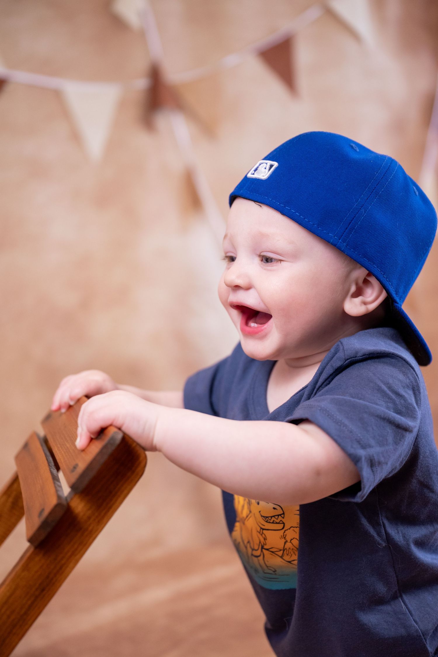 Happy toddler boy wearing a blue snapback cap laughing while holding a wooden chair railing.